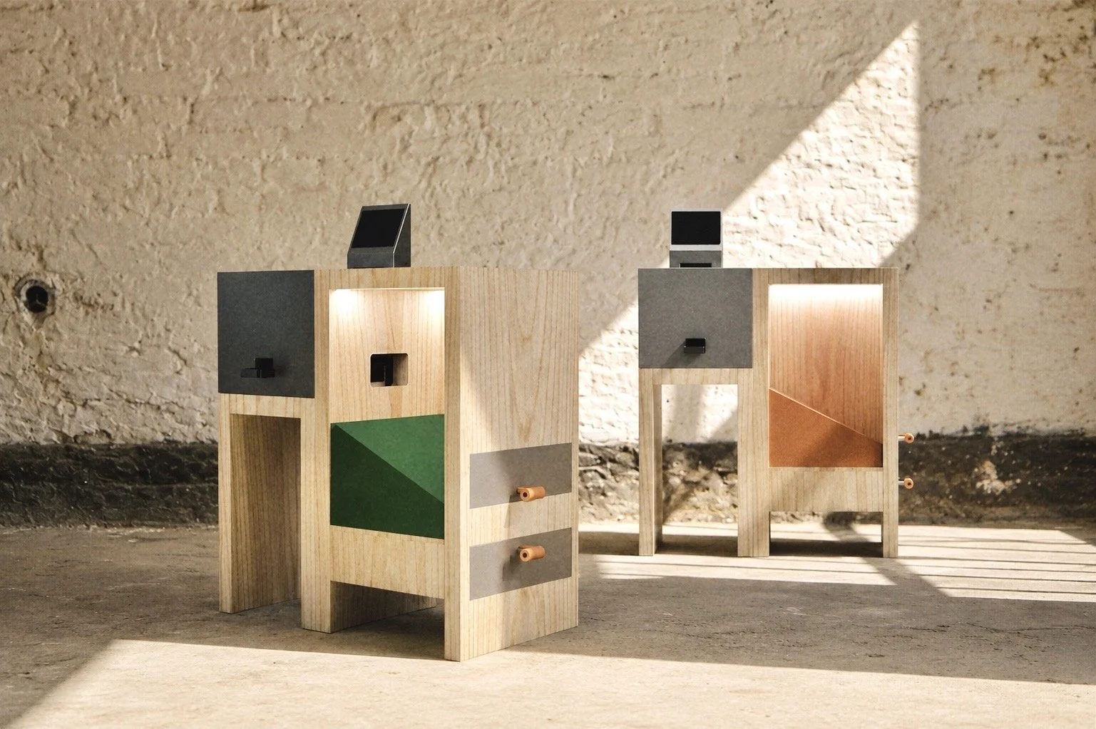 Two small wooden display tables with colorful geometric paper designs and small black screens on top, set against a textured brick wall with sunlight and shadow patterns.