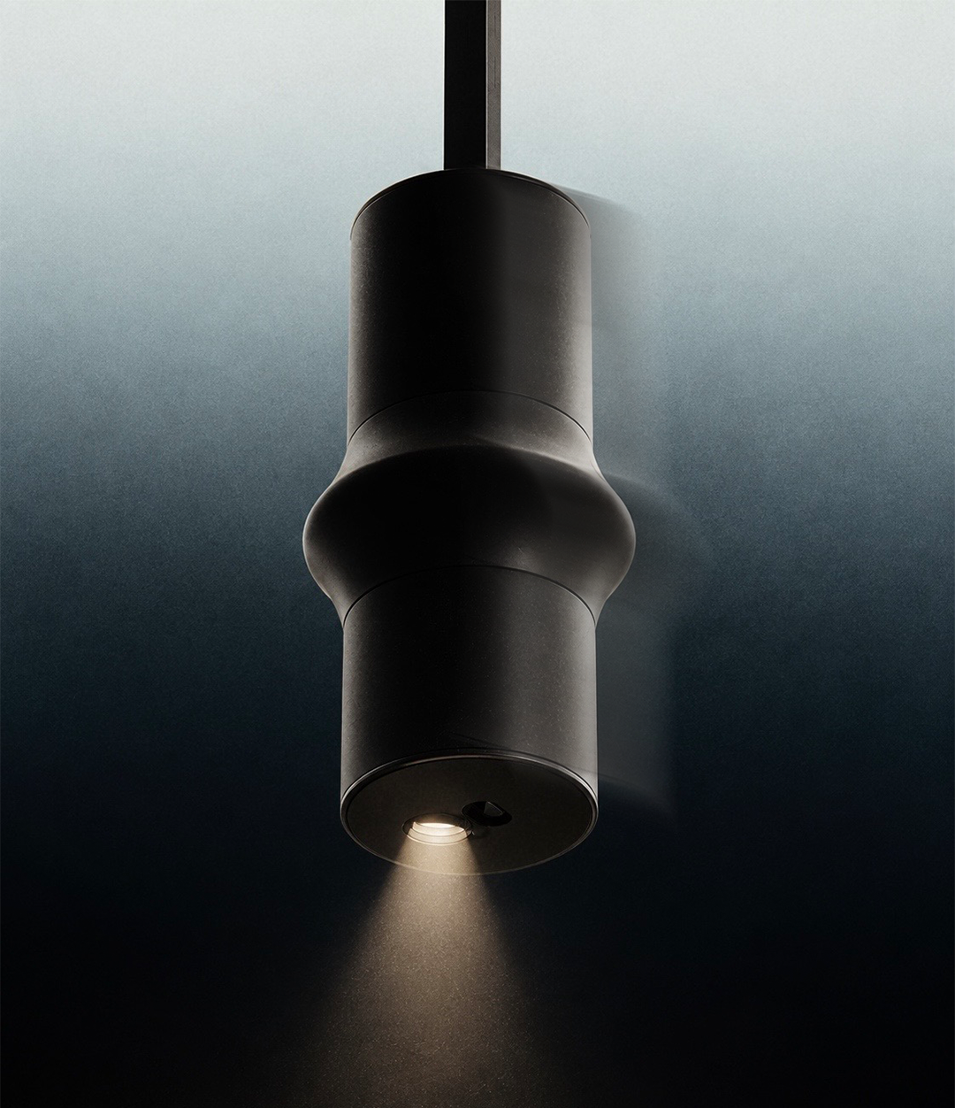 A black, modern ceiling spotlight illuminating downward, mounted on a ceiling with the light creating a focused beam.