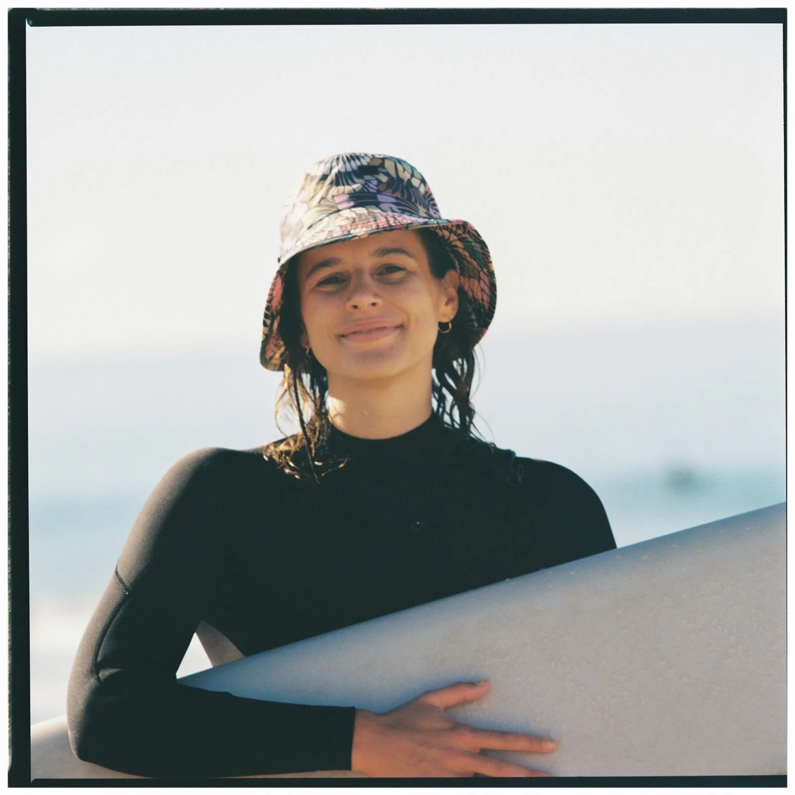 Frau steht mit nassen Haaren und mit Longboard vor dem Meer.