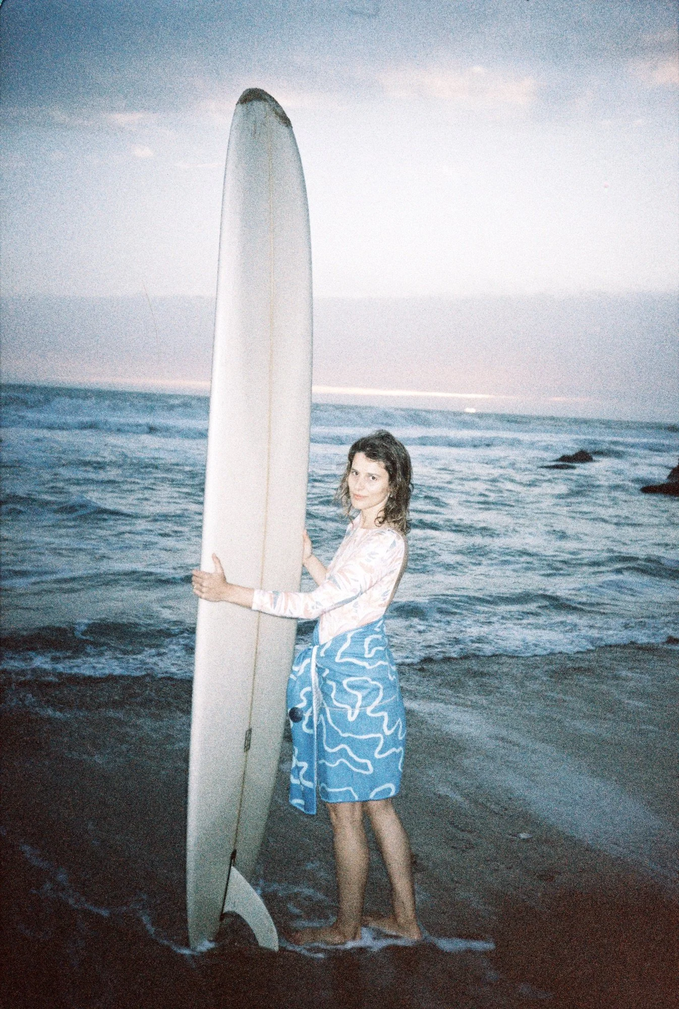 Frau steht im Meer am Strand, hält ein Surfbrett und trägt eine blaue Strandhose, die mit weißen Mustern bedeckt ist, bei Sonnenuntergang.
