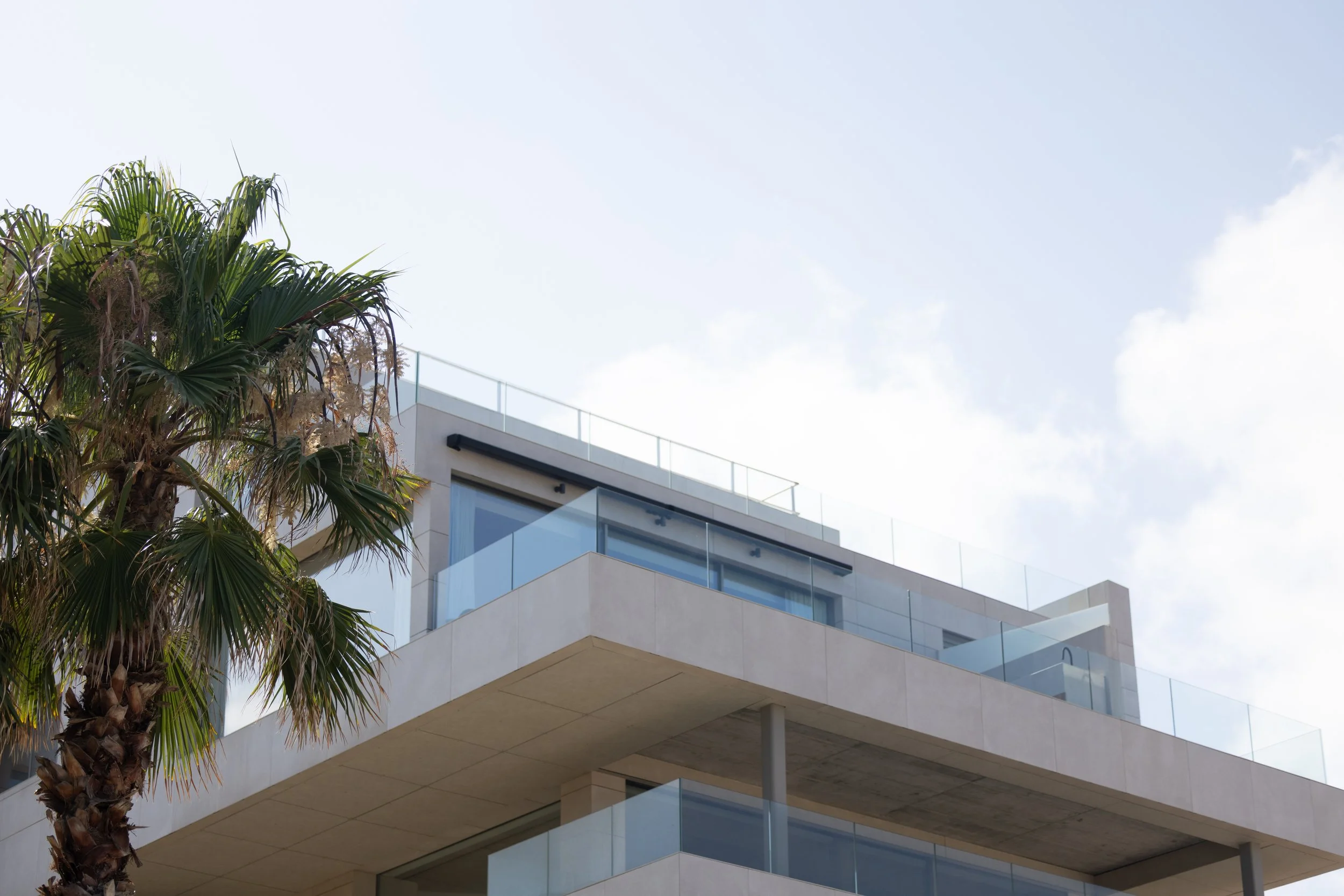 A modern multi-story building with glass railings and large windows, partially obscured by a palm tree, against a blue sky with some clouds.