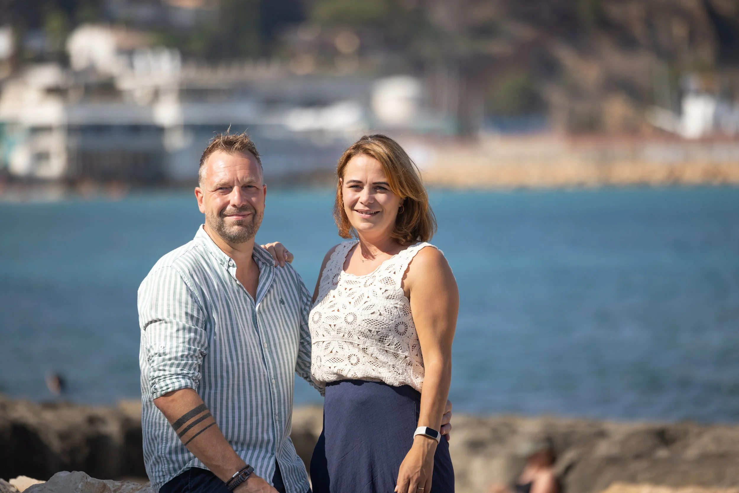 A man and woman standing outdoors near water, smiling at the camera, with blurred boats and buildings in the background.