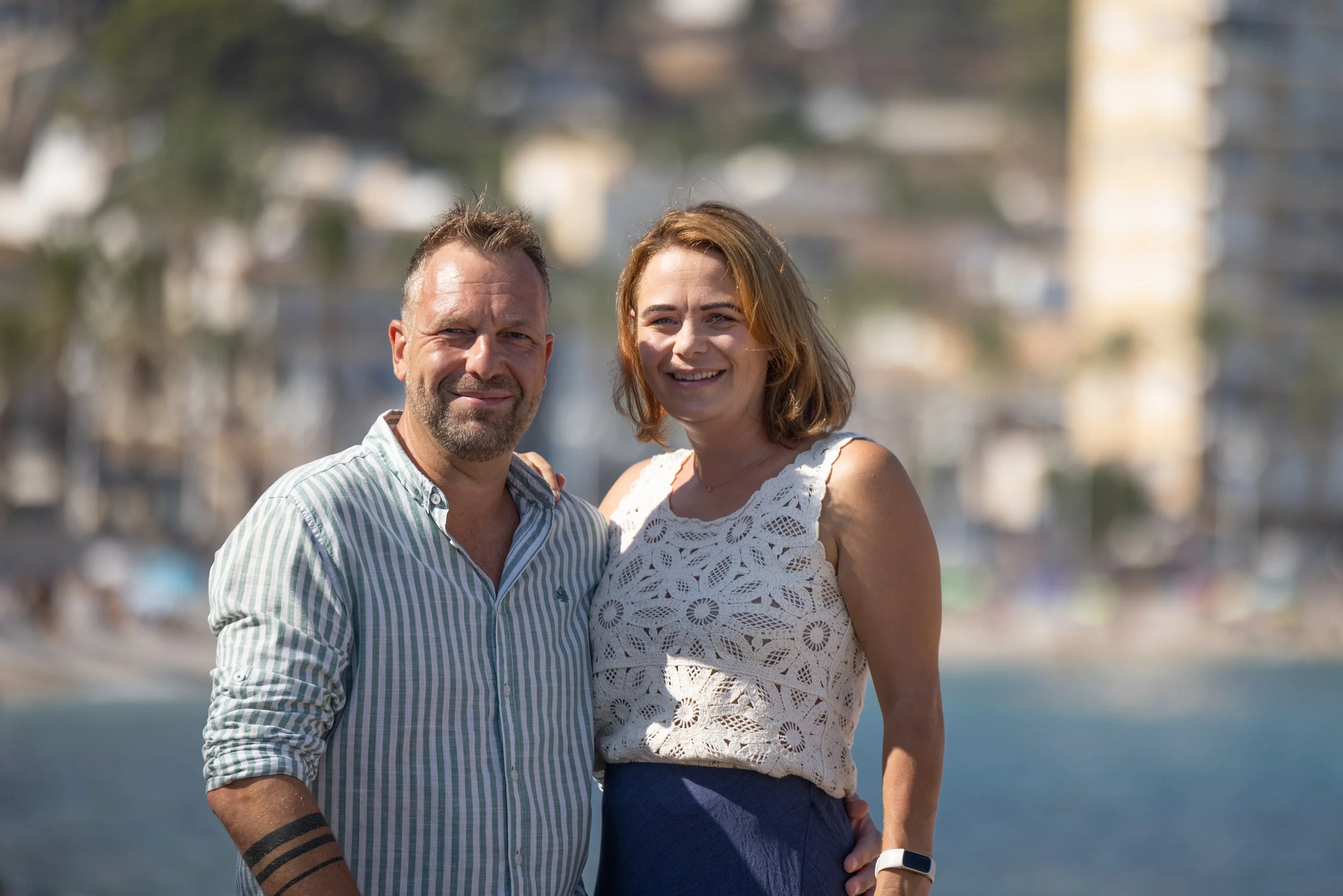 A man and woman smiling outdoors near water with buildings and trees in the background.