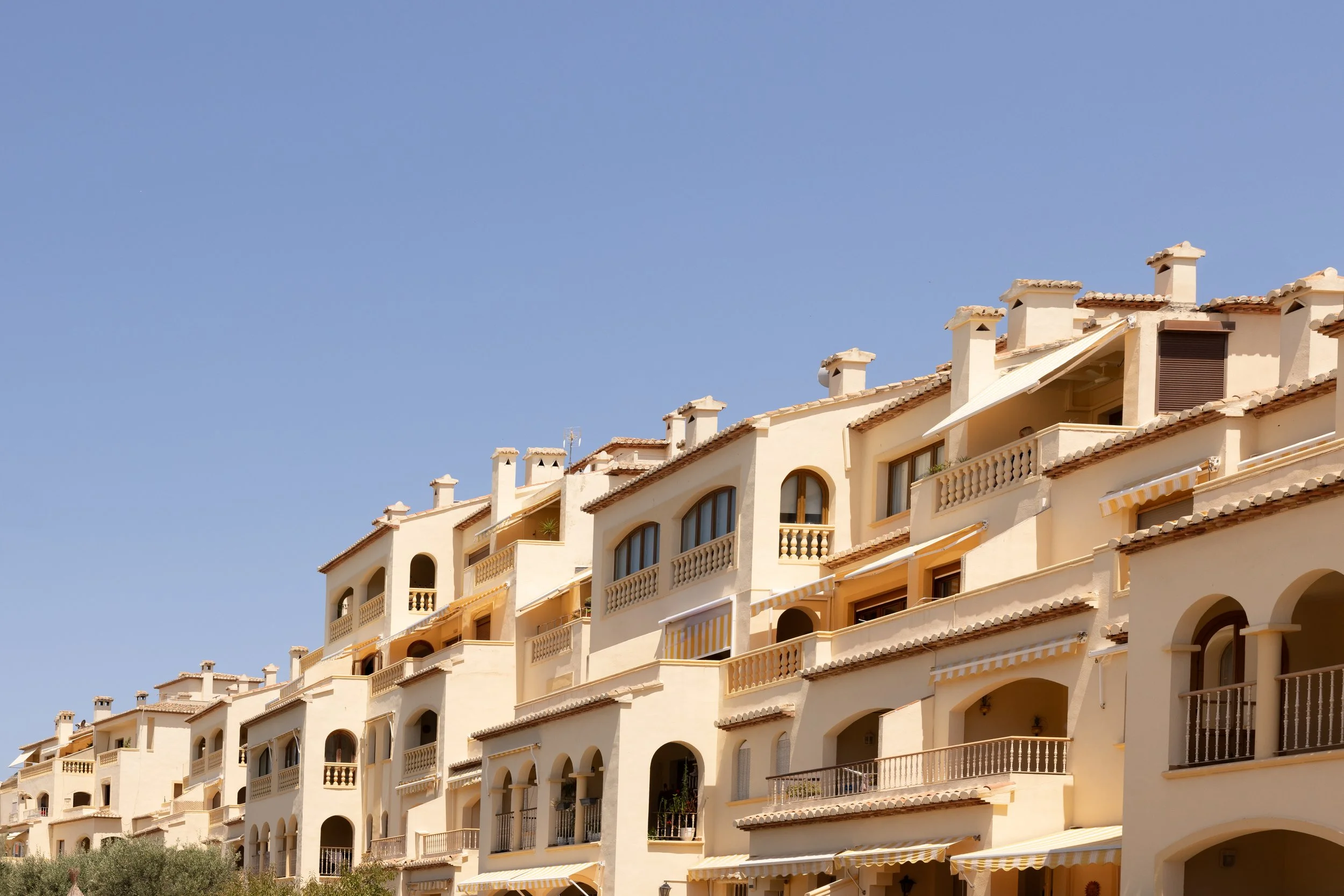 Multi-story Mediterranean-style apartment buildings with balconies and awnings under a clear blue sky.