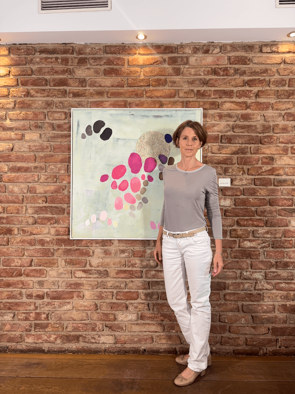 Artist Birgit Trofer at her exhibition with an artwork of the series Dancing Palette mounted on a brick wall in a historical building.
