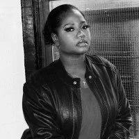 Black and white photo of a young woman with her hair in a bun, wearing a leather jacket, standing in front of a textured background.