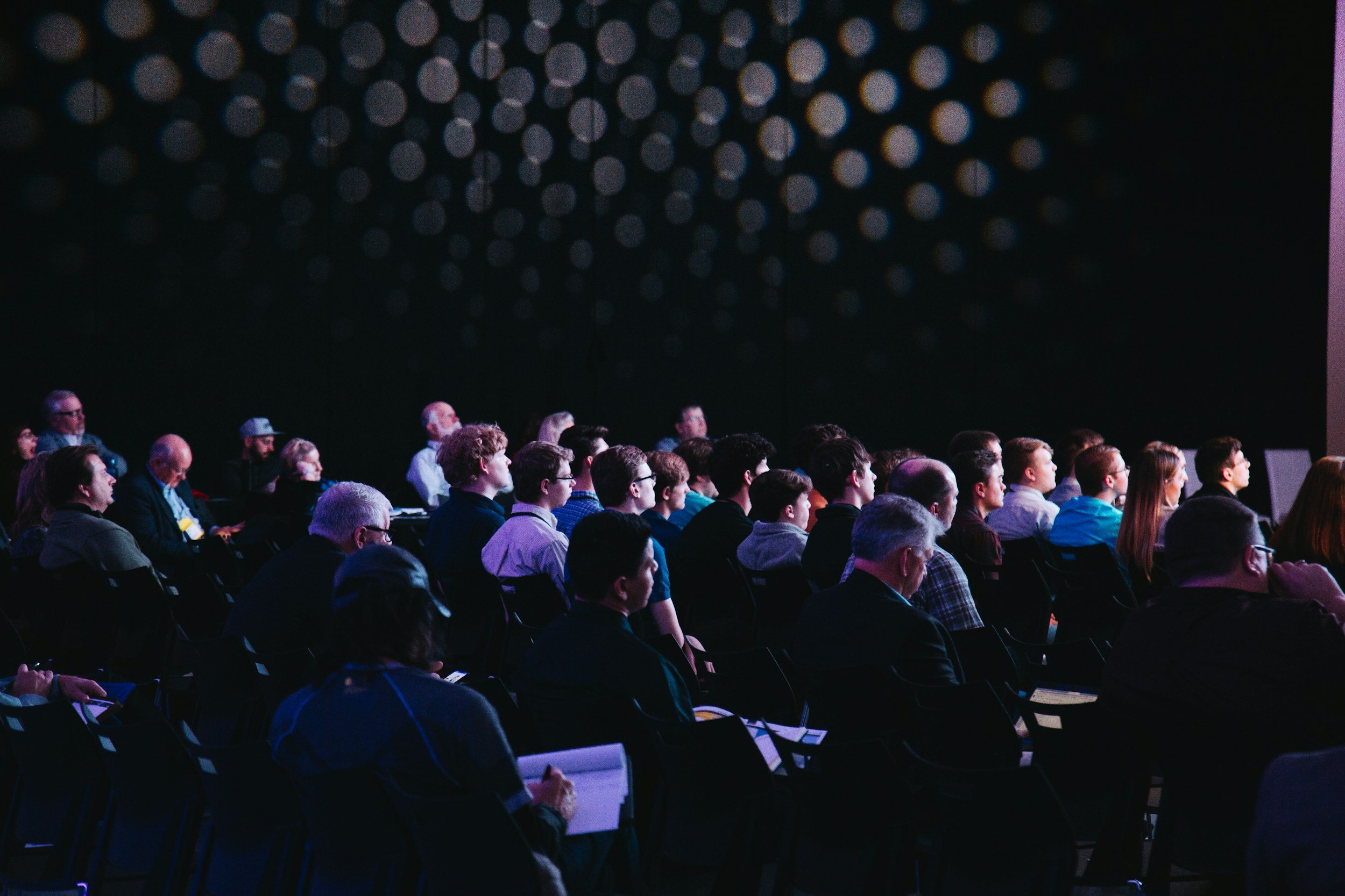 Audience of diverse adults seated and attentively watching a presentation or speaker at a conference in a dark room with decorative circular light pattern on the wall.