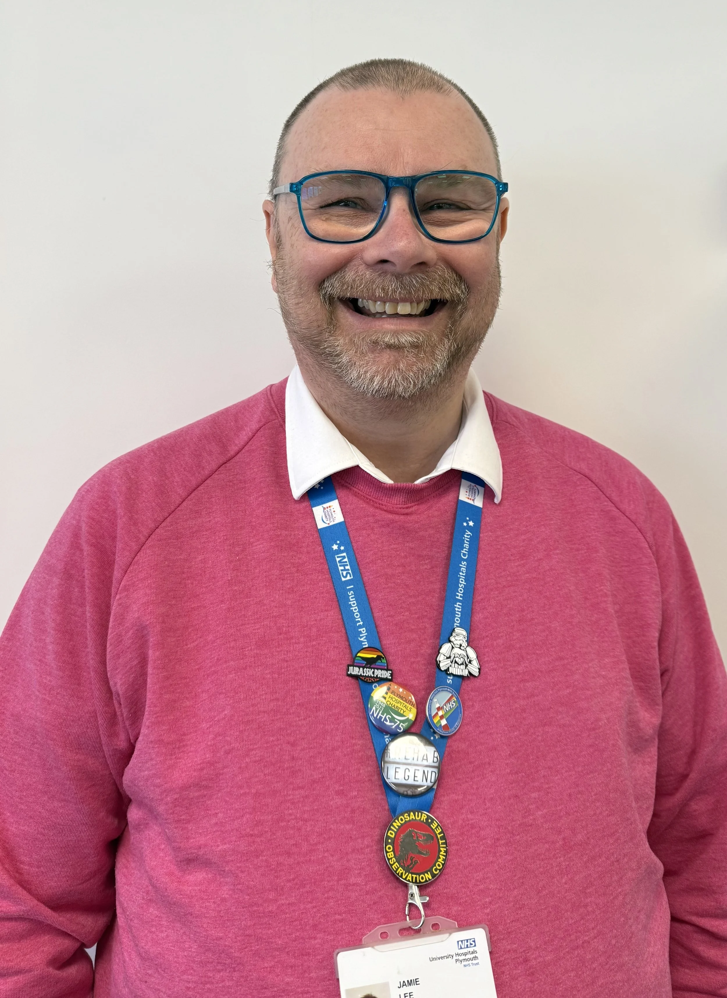 A smiling man with glasses, a beard, and a buzz cut, wearing a pink sweater over a white collared shirt, with a blue lanyard and multiple pins, standing against a plain white background.
