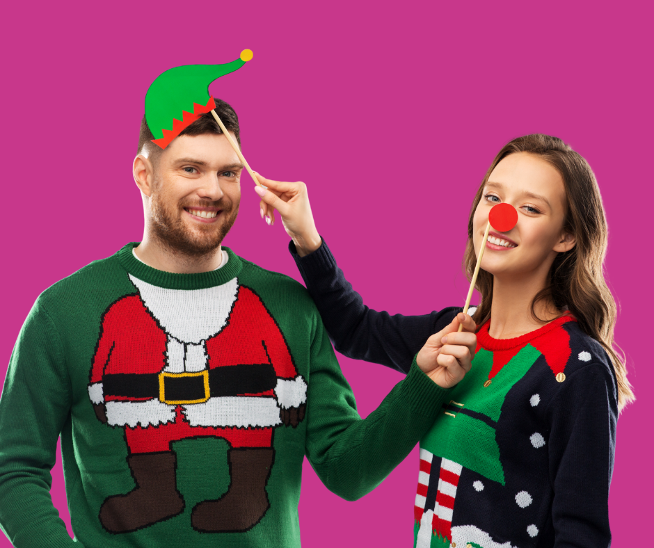 A man and woman in festive sweaters playfully hold props; the man has a green elf hat, the woman a red nose. They smile against a pink background, creating a joyful holiday mood.