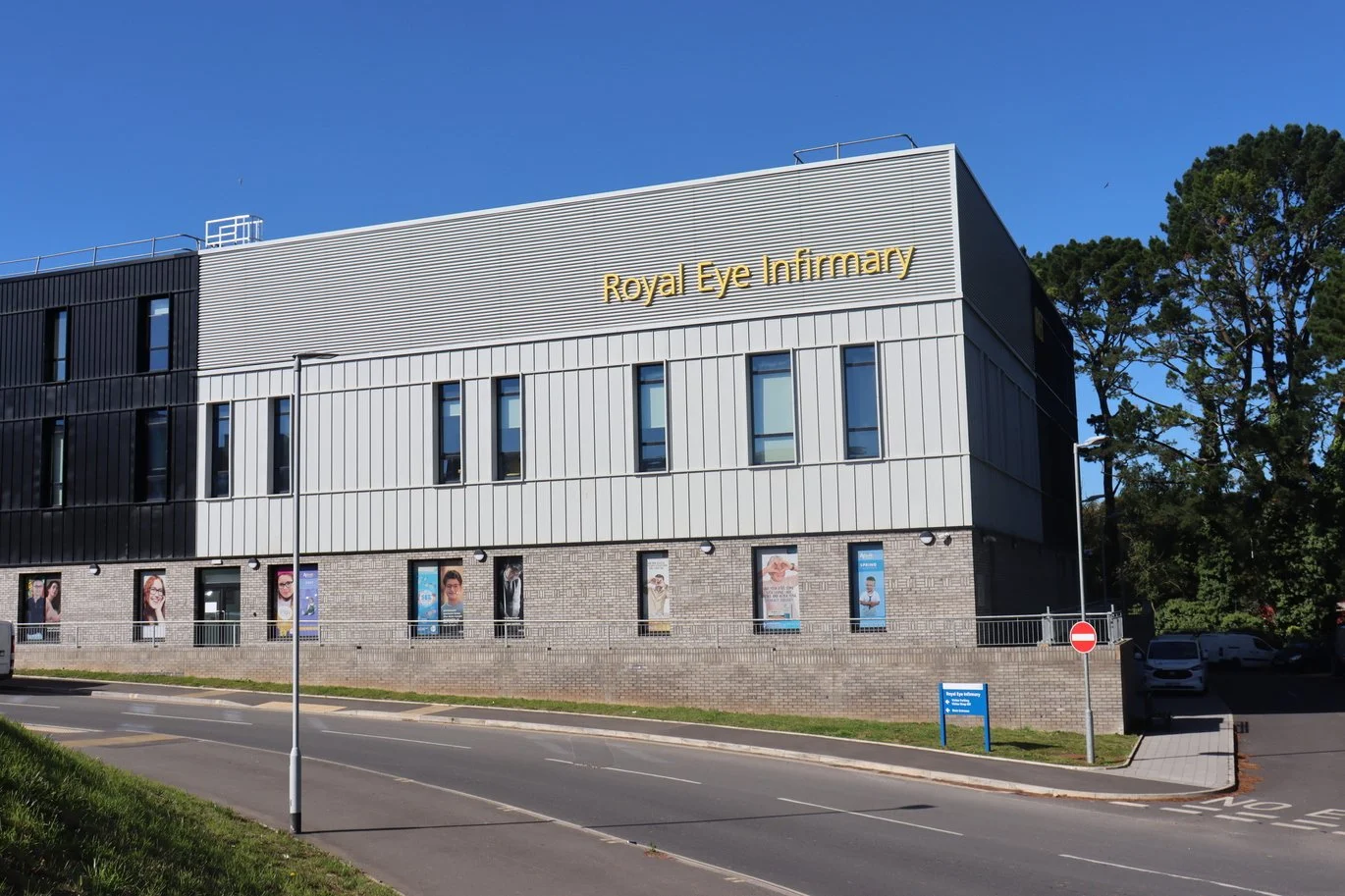 Exterior view of the Royal Eye Infirmary building with a modern facade, multiple windows, and posters on the lower level, situated beside a road with trees in the background.