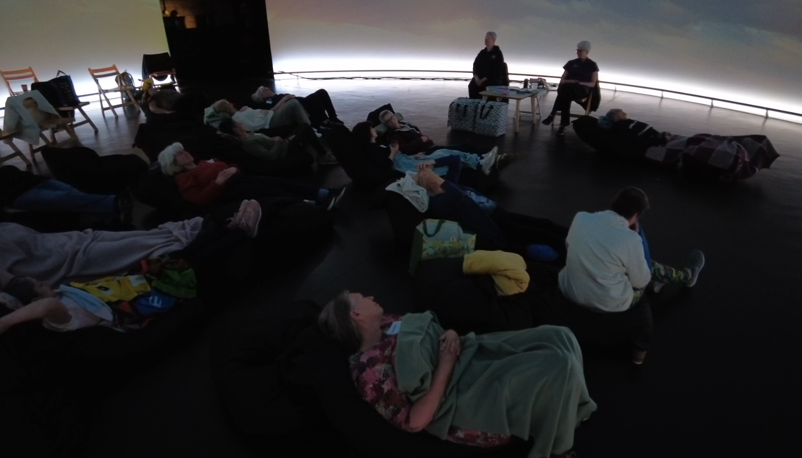 In a dark room, workshop attendees lie on bean bags under blankets, gazing up at an immersive dome filled with soft, drifting clouds.
