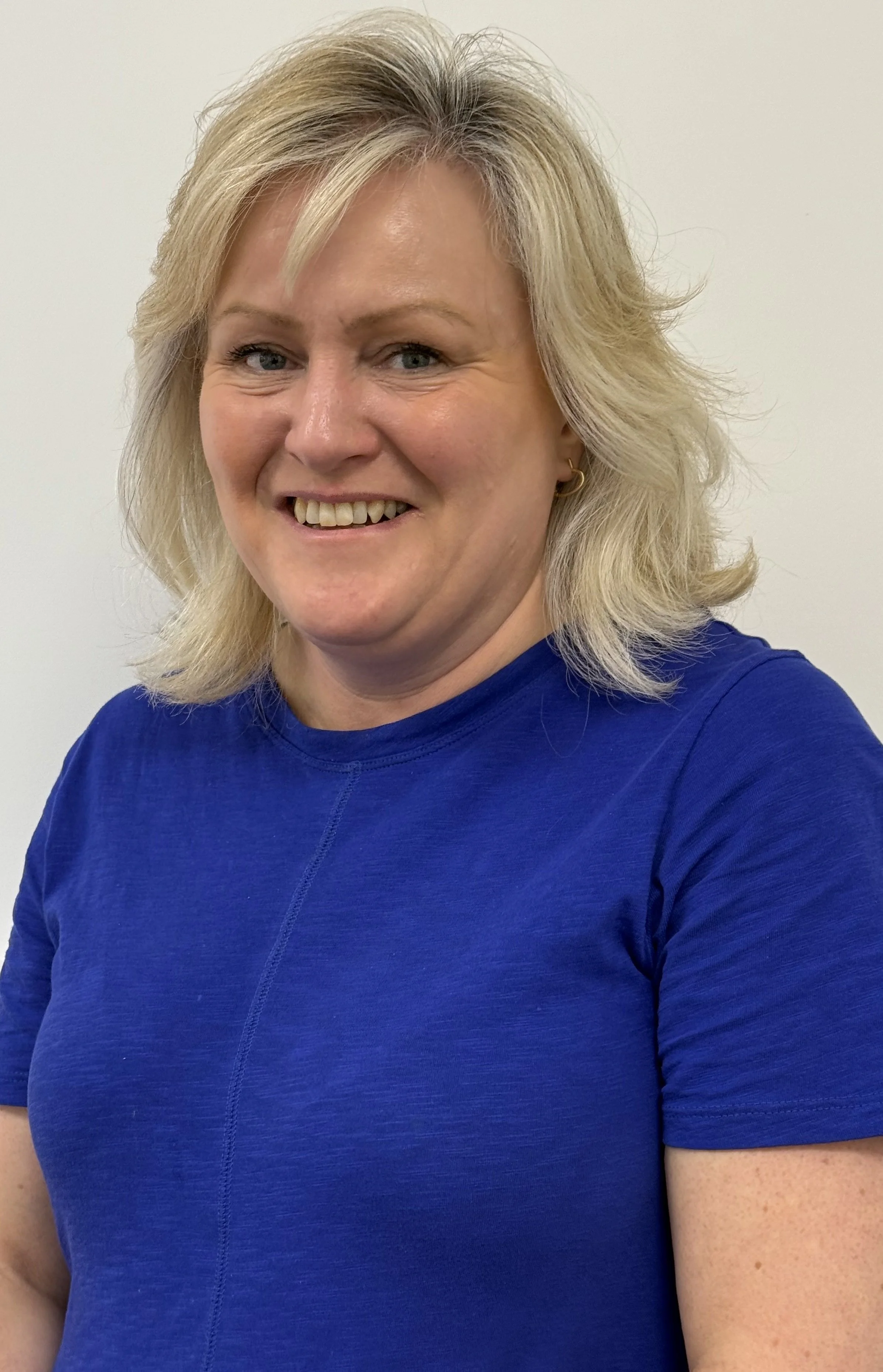 A smiling woman with blonde hair wearing a blue shirt against a plain white background.