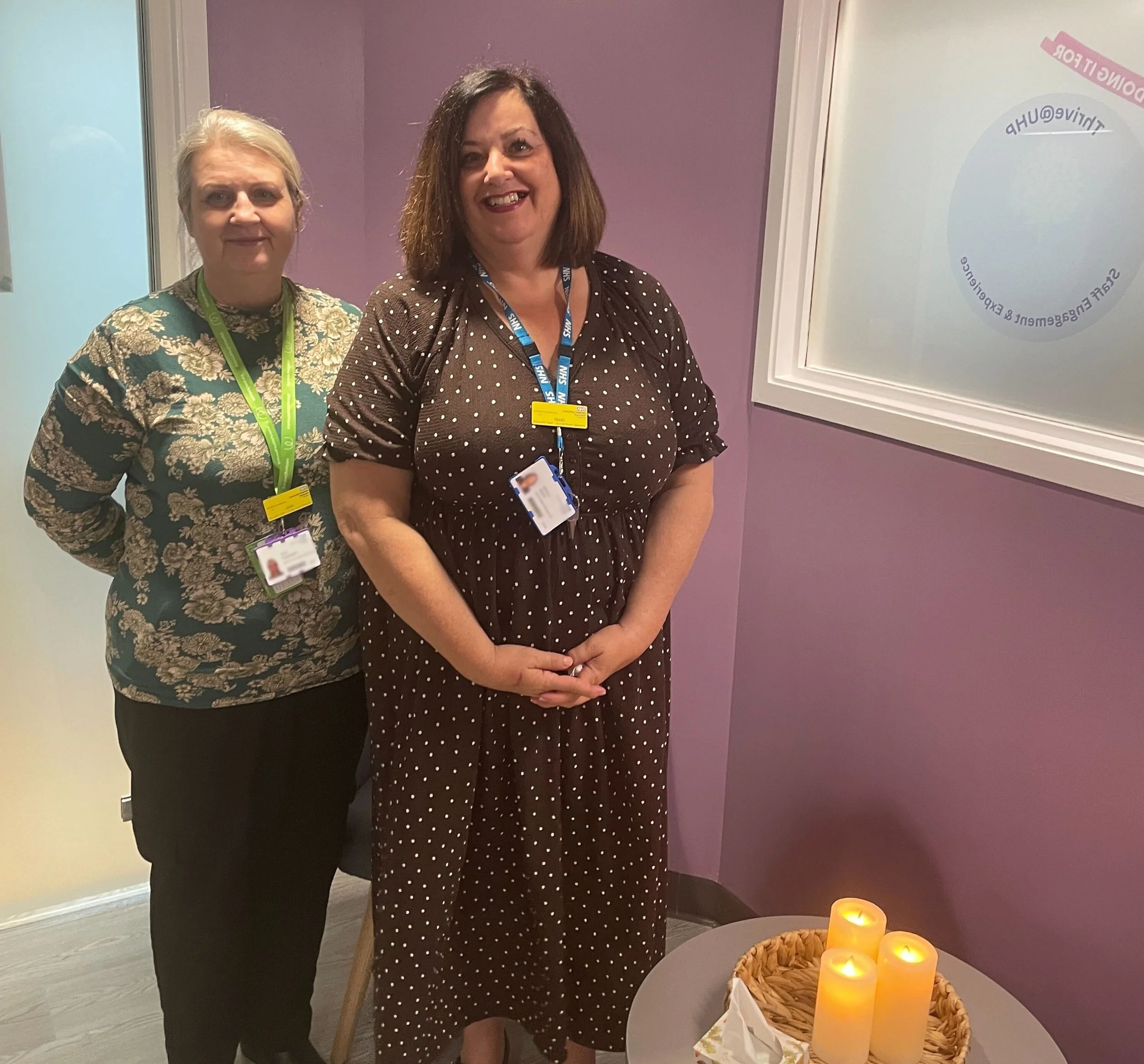 Nikki and Julie, End of Life Educators, smiling for a photo. They are stood next to a small table with led pillar candles and a box of tissues.
