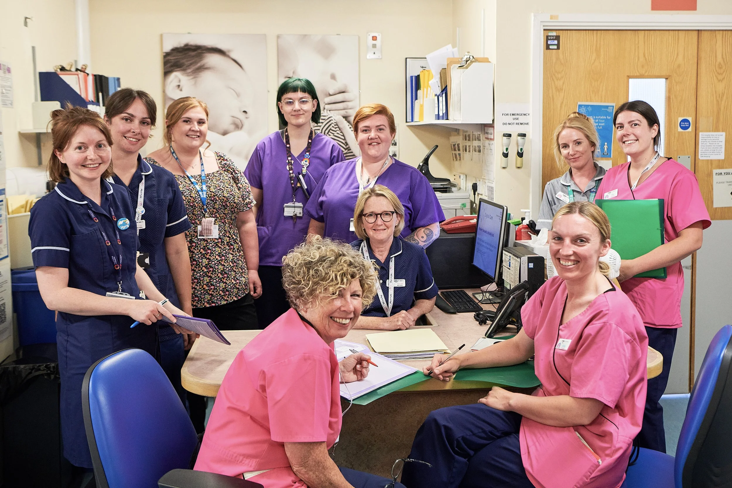 Group of healthcare professionals in a hospital room, posing for a photo at a desk with computers and medical supplies, smiling and wearing scrubs of various colors.