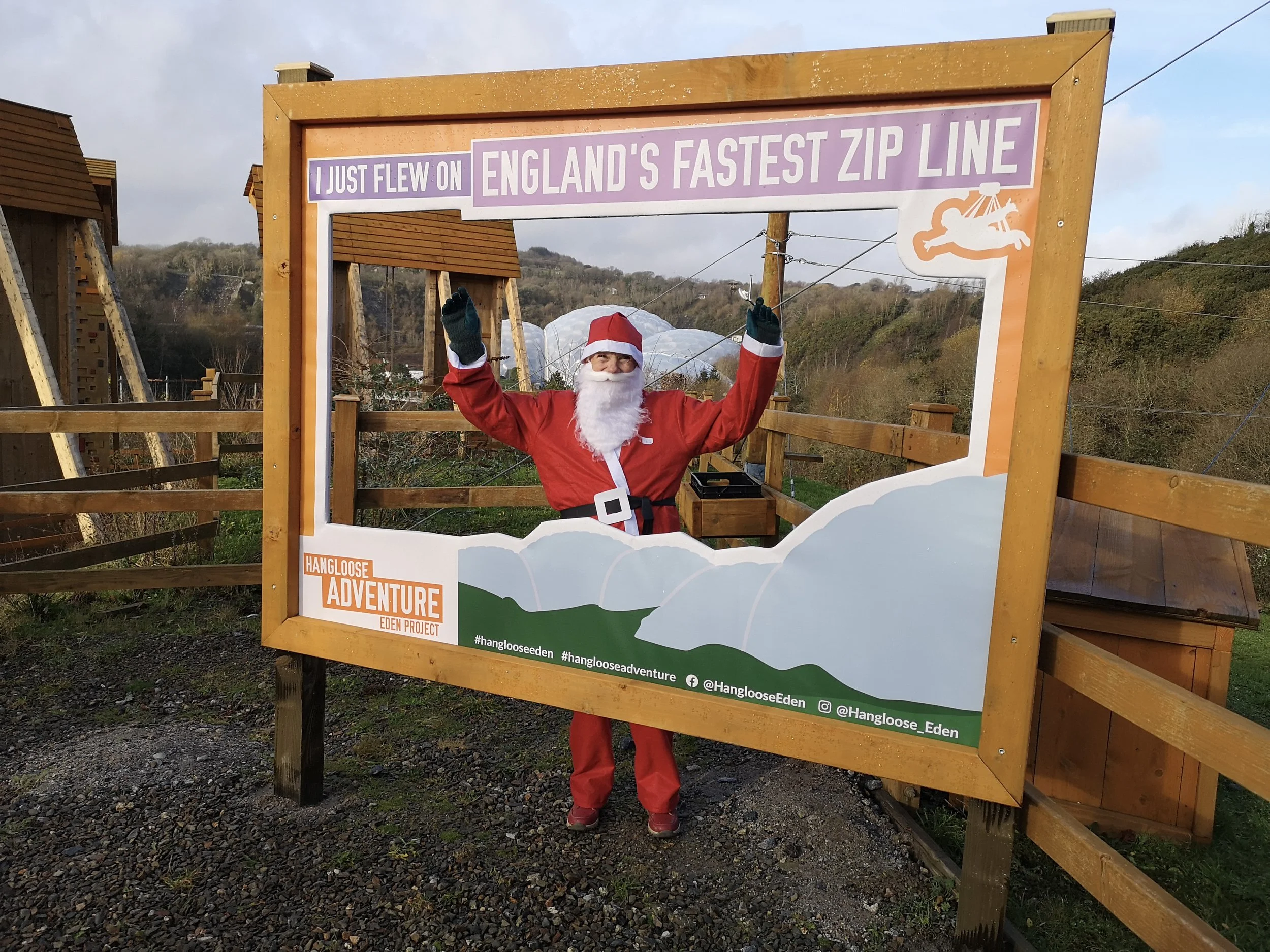 Person dressed as Santa Claus with arms raised, standing behind a wooden frame that says 'I just flew on England's fastest zip line', during daytime with hills and a cloudy sky in the background.