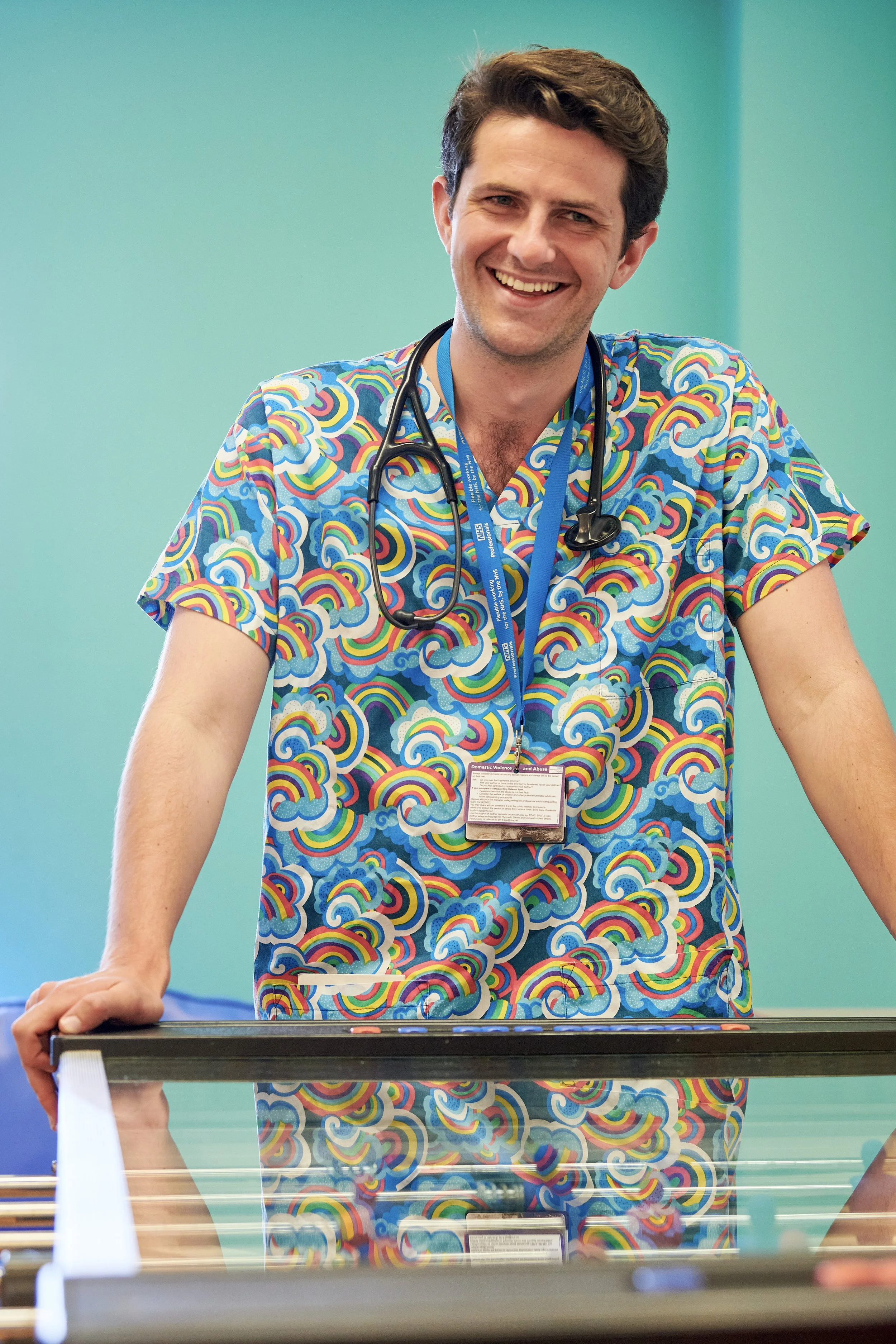 A young male doctor smiling, wearing colorful scrubs with rainbow cloud patterns, standing behind a reflective glass table in a medical setting with a turquoise wall.