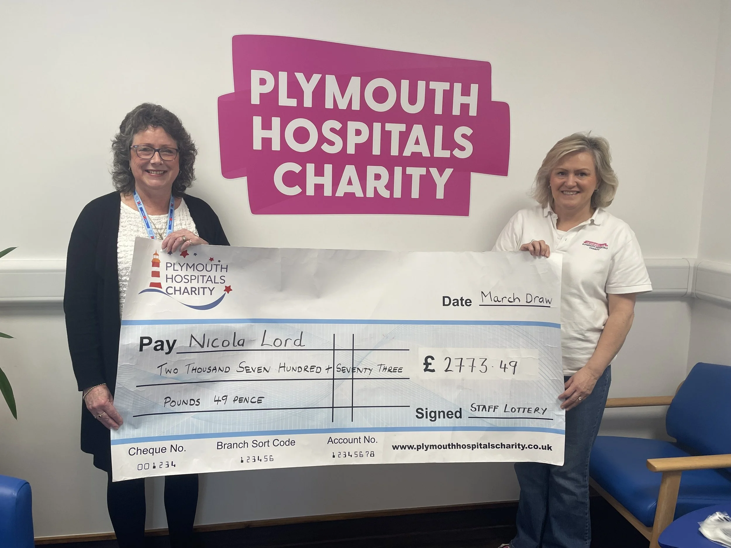 Nicola holding a giant cheque beside Bernie, in front of a Plymouth Hospitals Charity logo