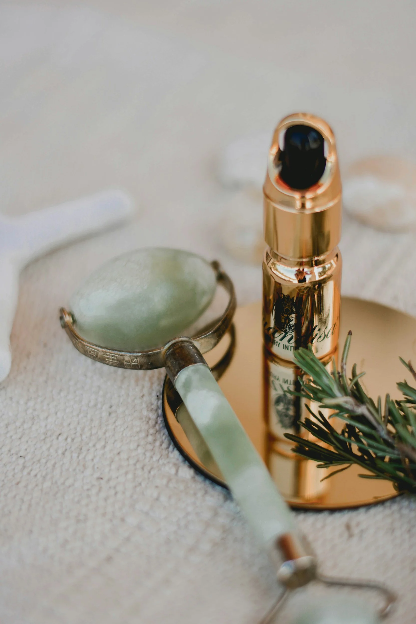 A jade facial roller, a bottle of facial serum, and some greenery on a reflective surface.