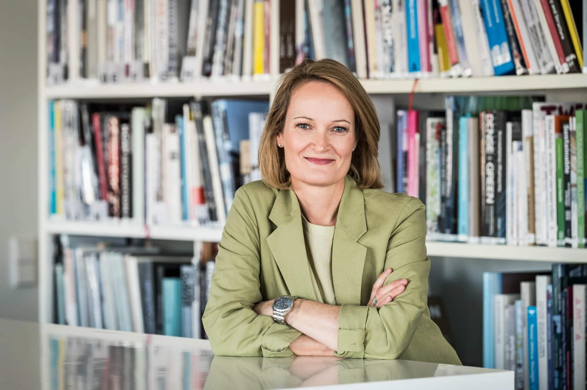 Eine Frau sitzt an einem Tisch in einer Bibliothek, umgeben von Büchern, trägt eine grüne Jacke und lächelt.