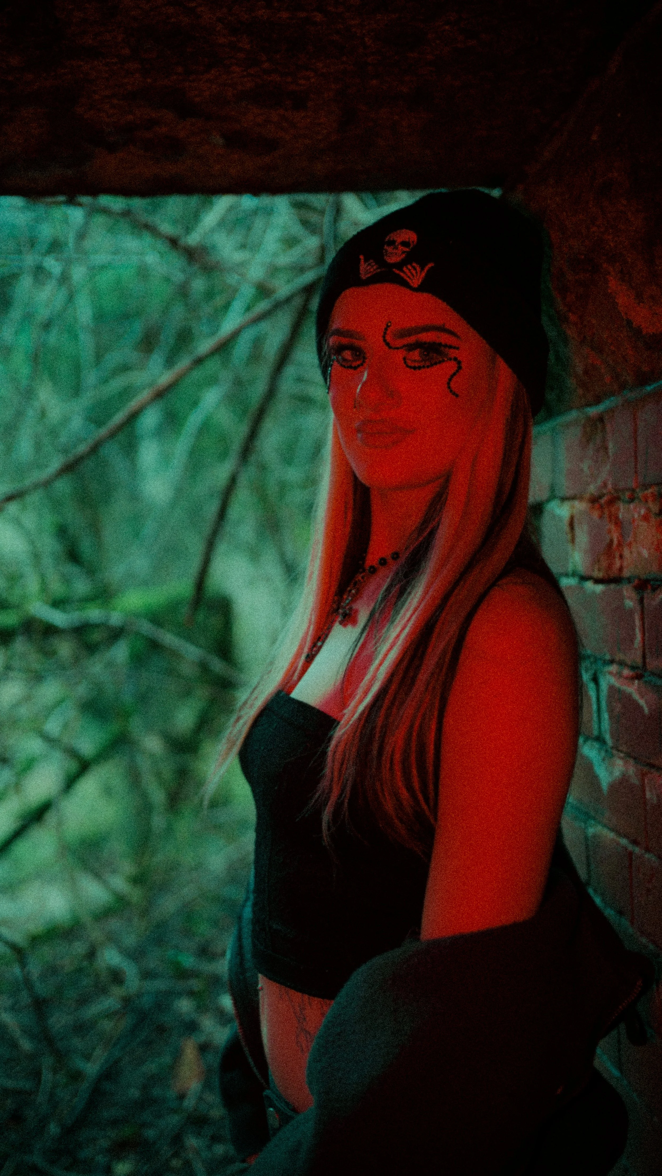 A woman with long blonde hair wearing a black beanie with a skull and crossbones, a black top, necklaces, and a jacket, standing next to a brick wall in a shaded outdoor area.