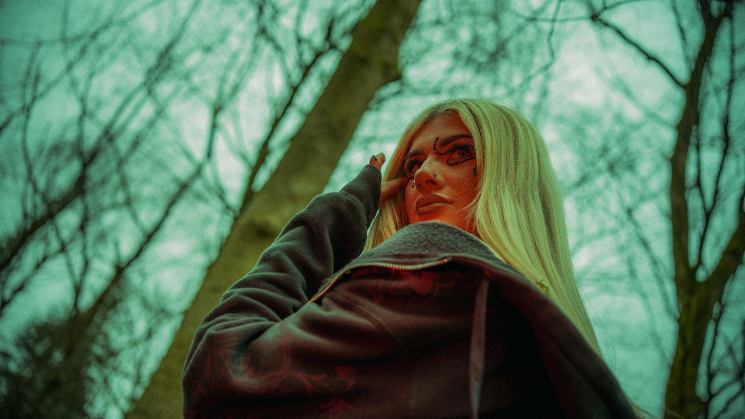 Young woman with bright blonde hair and makeup in a forest with leafless trees, looking upwards.