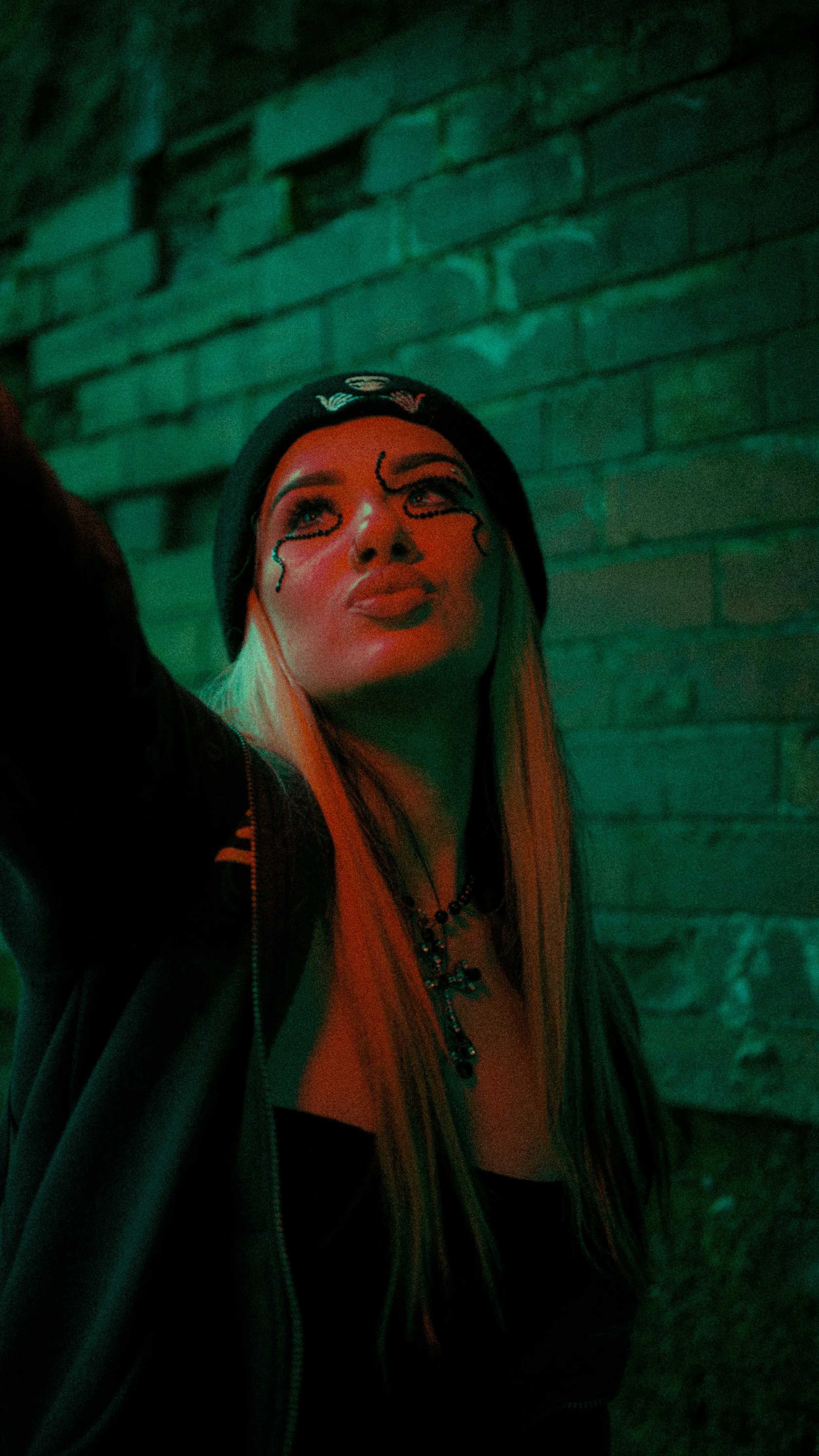 Woman with tattoos on face, long blonde hair, wearing a black beanie, black jacket, and necklace, standing against a green-lit brick wall.