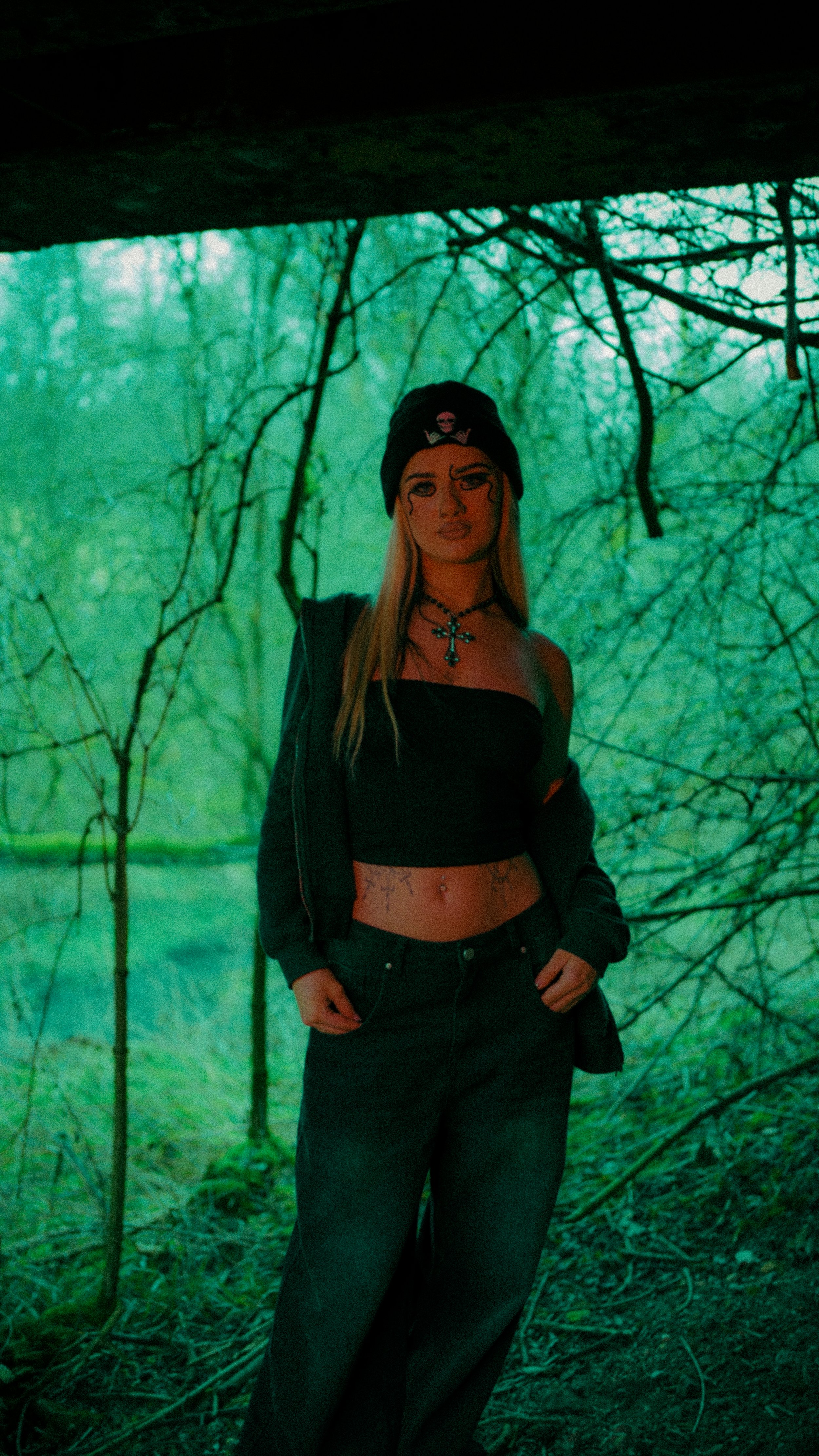 A woman standing in a wooded area with a background of green trees. She's wearing a black beanie with a pink skull, a black tube top, a black jacket, black pants, and accessories including a cross necklace and tattoos.