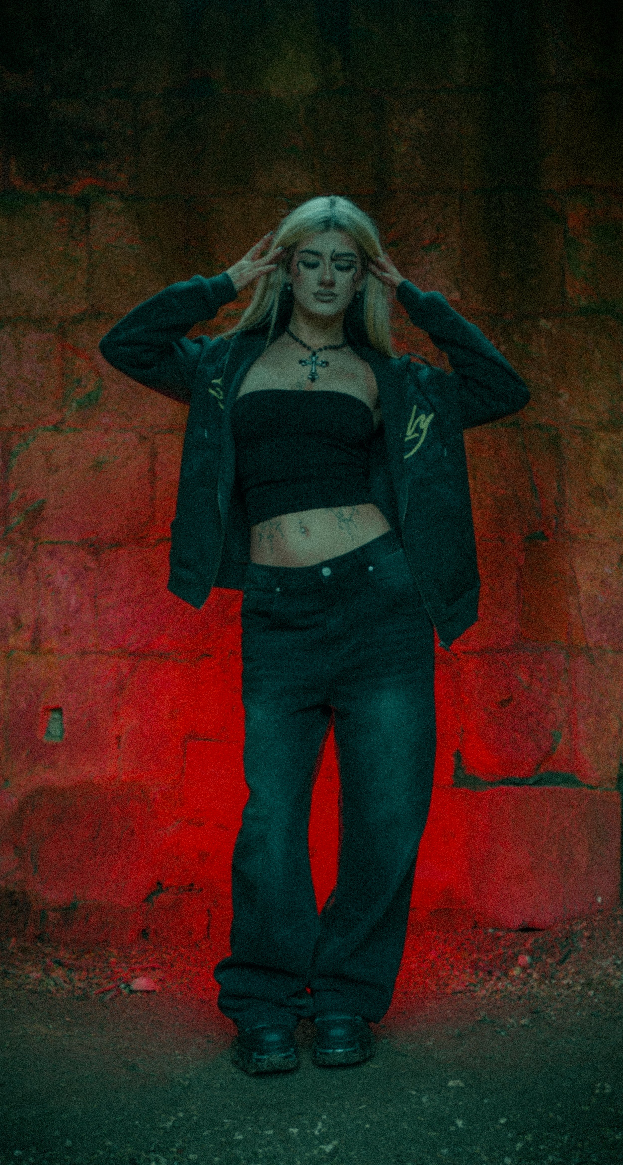 A young woman with long blonde hair standing against a red brick wall, wearing a black strapless top, black jeans, a black jacket, and black shoes, with tattoos visible on her midriff and chest.