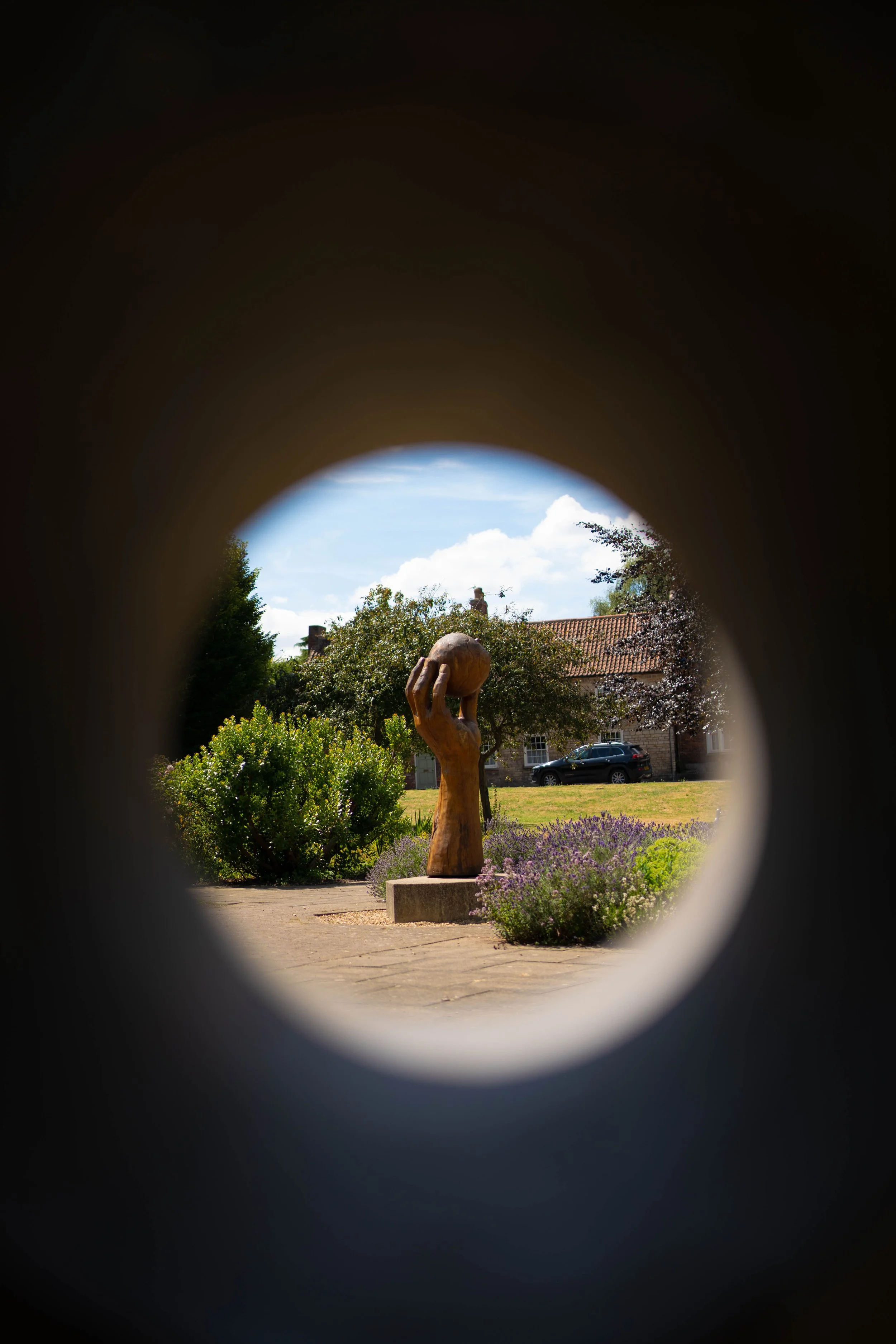 A sculpture of a hand holding a globe, viewed through a circular opening, outdoors with greenery, flowers, and a house in the background.