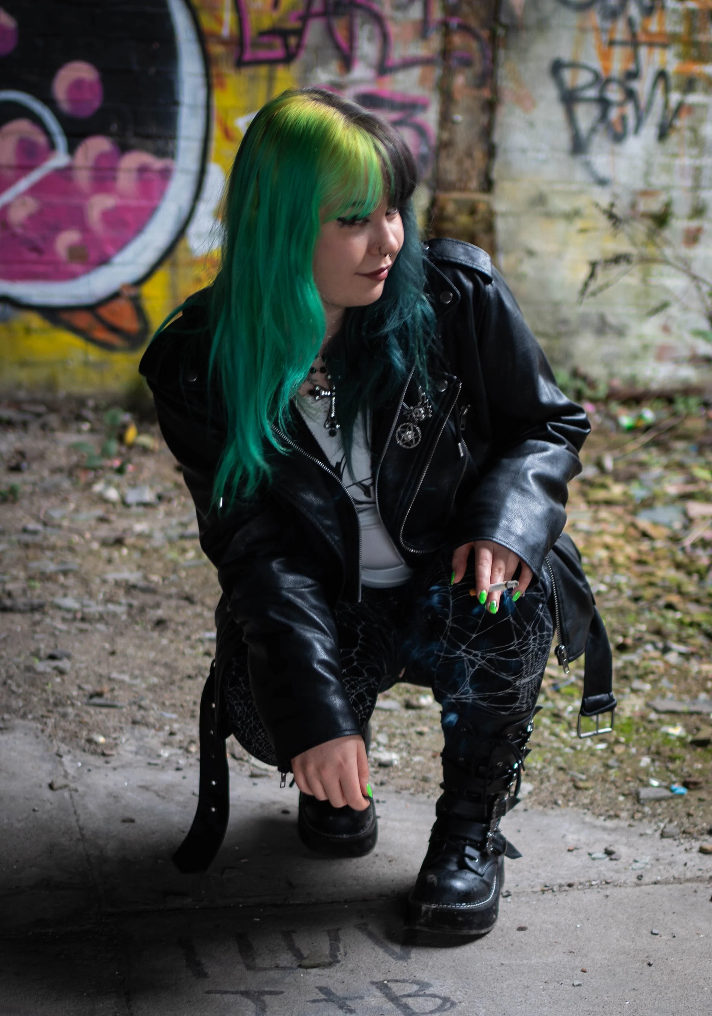A woman with colorful green and blue hair, wearing a black leather jacket and studded pants, crouching in front of a graffiti-covered brick wall in an urban setting.