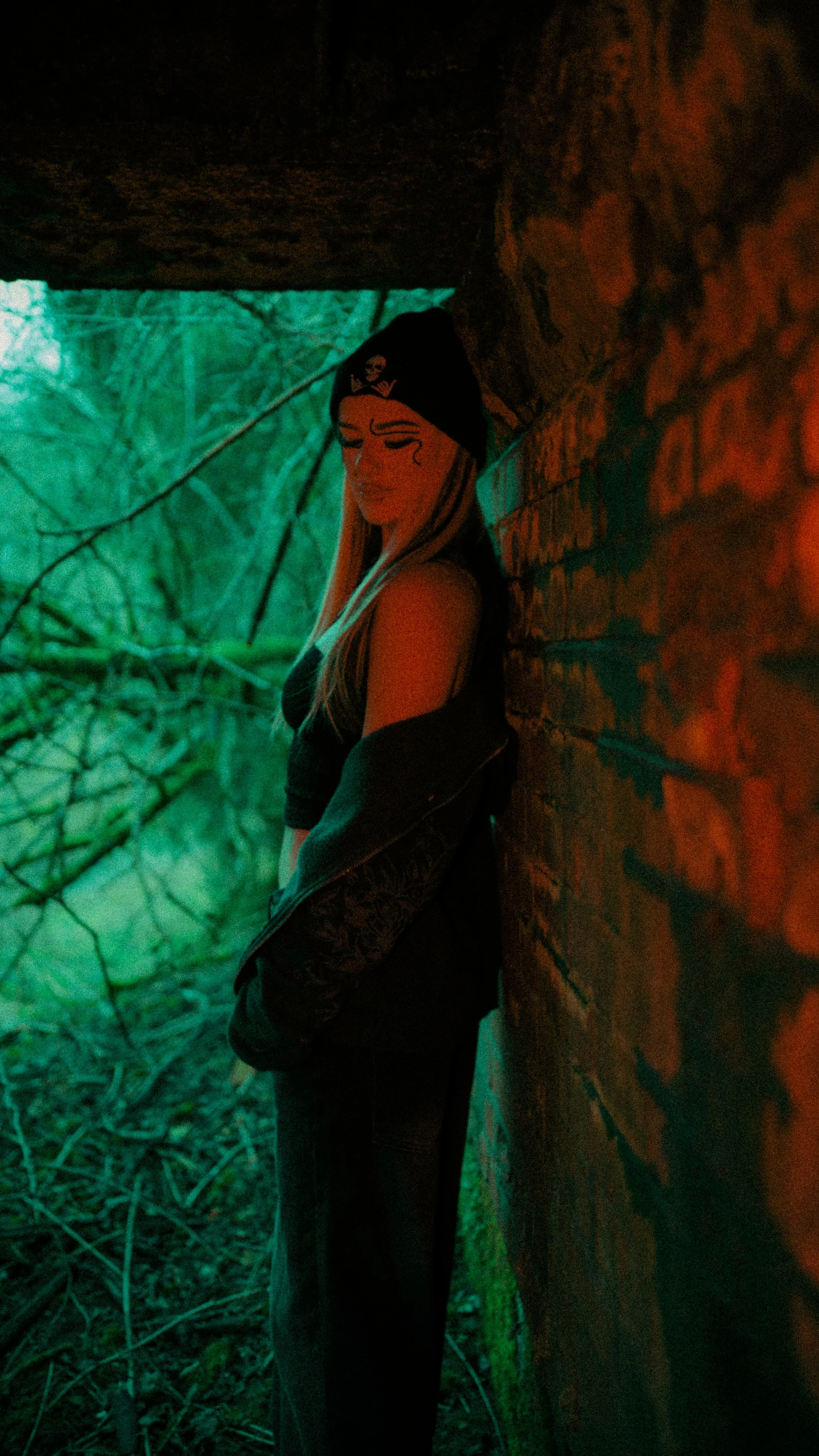A person with long hair wearing a black beanie with a skull and crossbones, black sleeveless top, and black pants, leaning against a brick wall in a dark outdoor area with green foliage in the background.