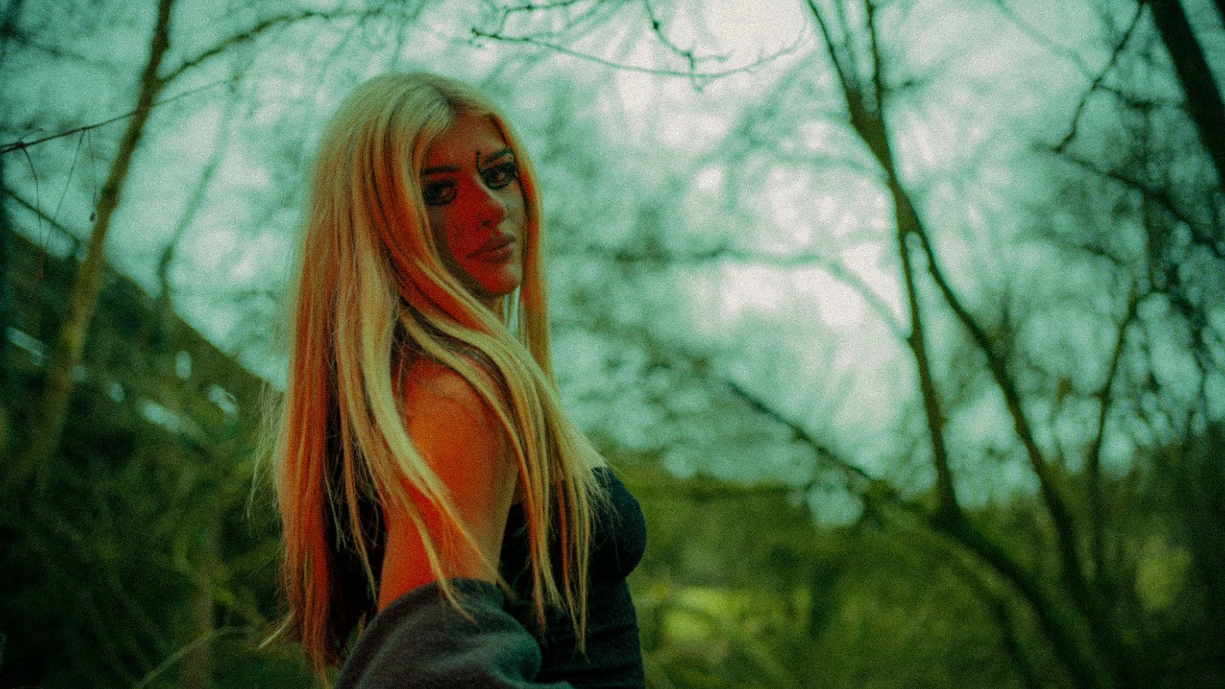 A woman with long blonde hair and drawn face makeup stands in a forest with green light filtering through the trees.