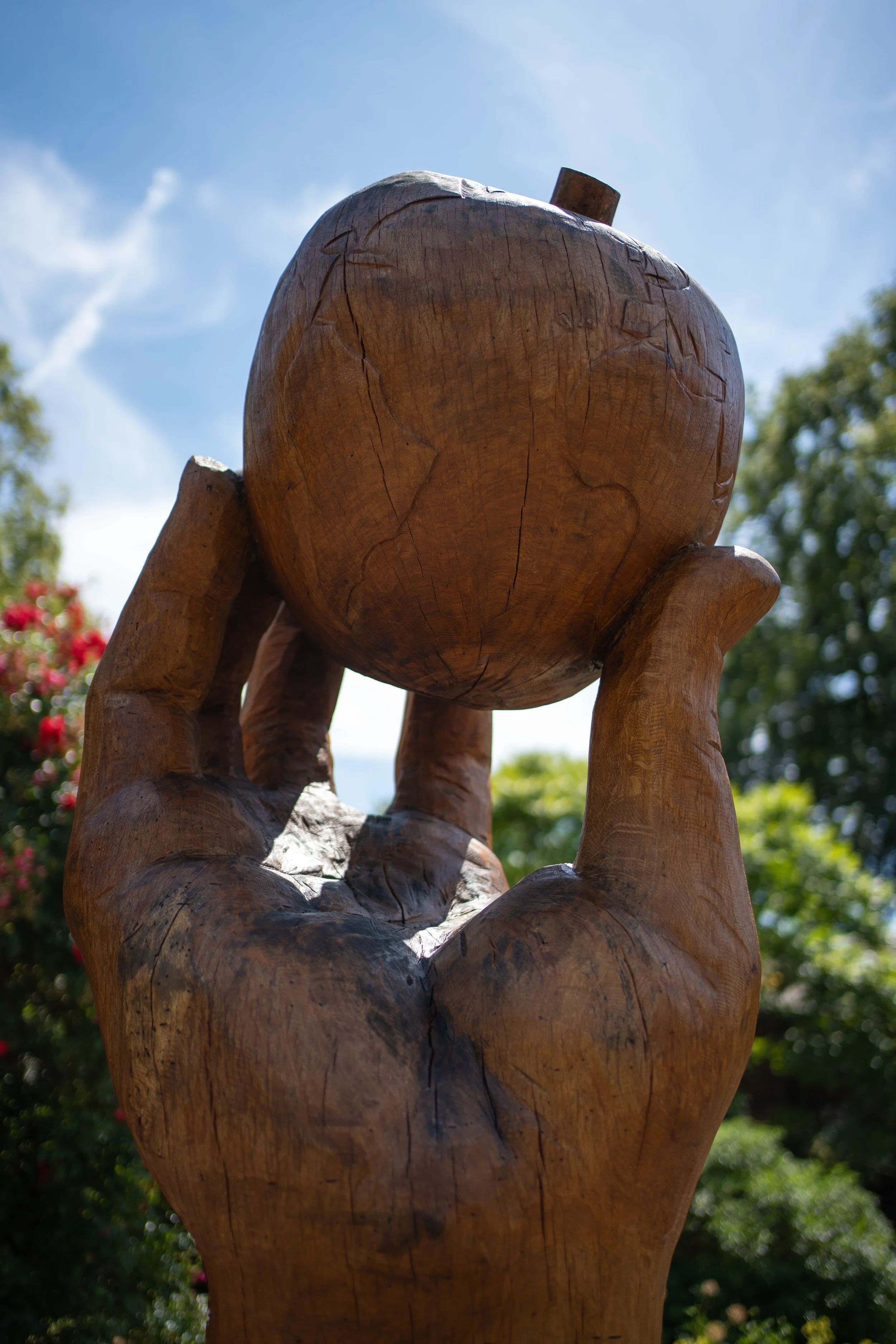 A wooden sculpture of a hand holding a globe, outdoors on a sunny day with a blue sky and trees in the background.