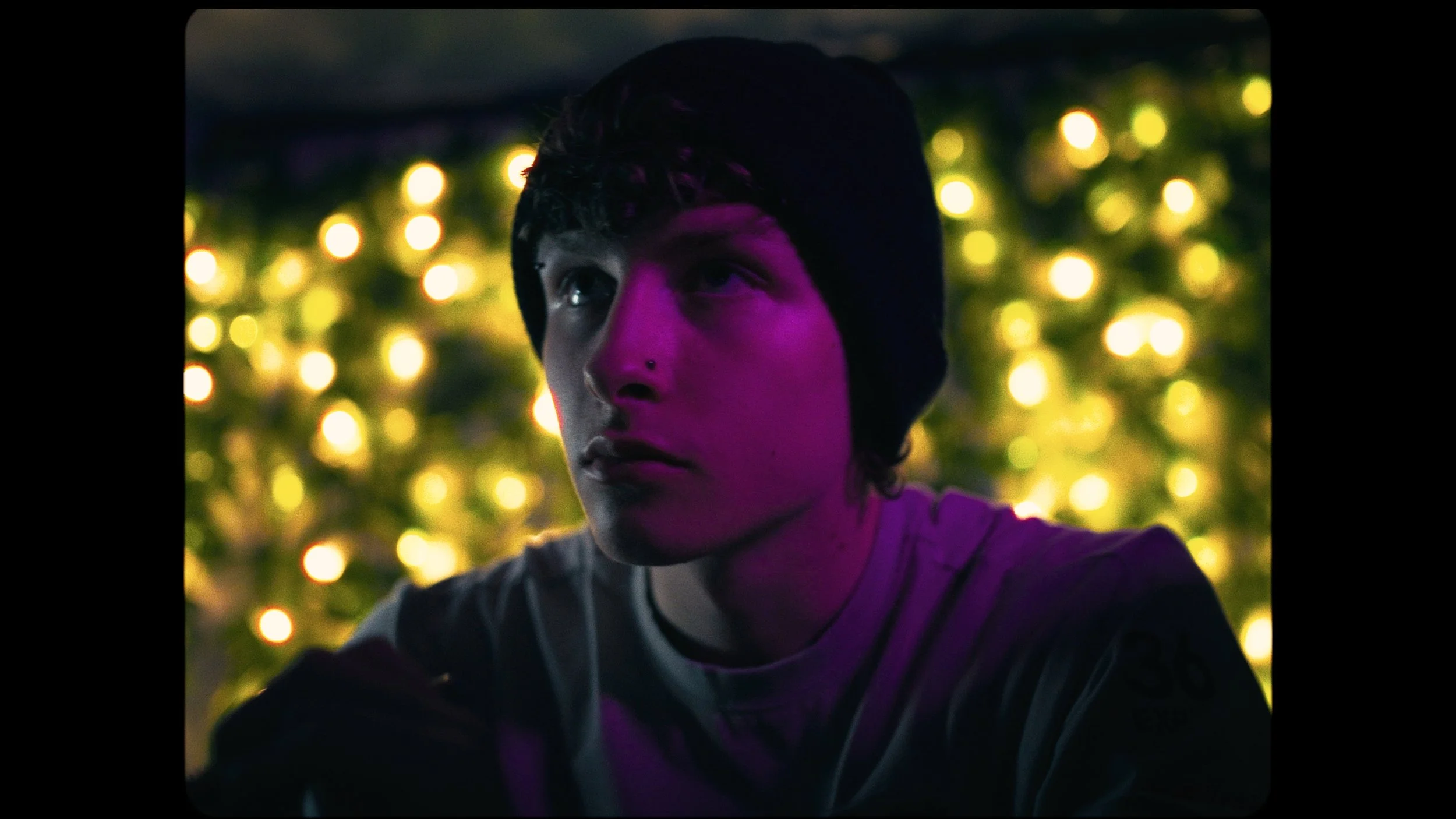 A young person with a nose piercing and a beanie, illuminated by purple lighting, with a blurred background of yellow lights.