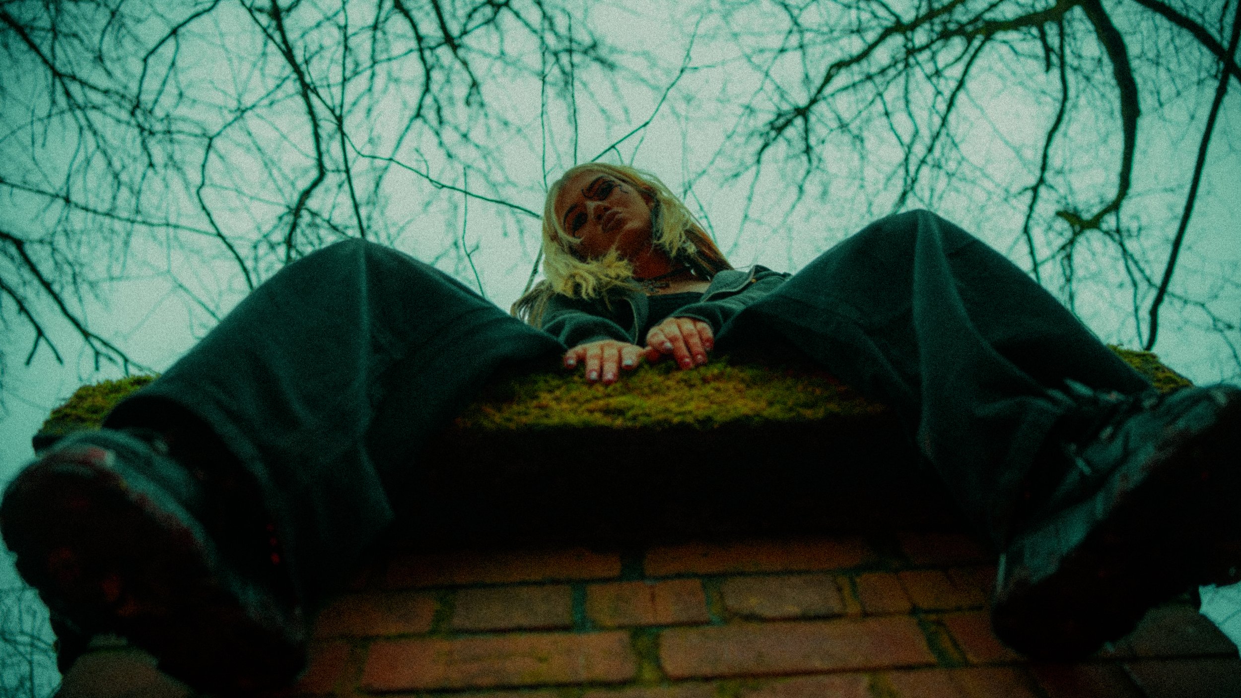 A girl with blonde hair and glasses leaning over a brick chimney with her hands resting on the edge, looking downward, with leafless trees and an overcast sky in the background.