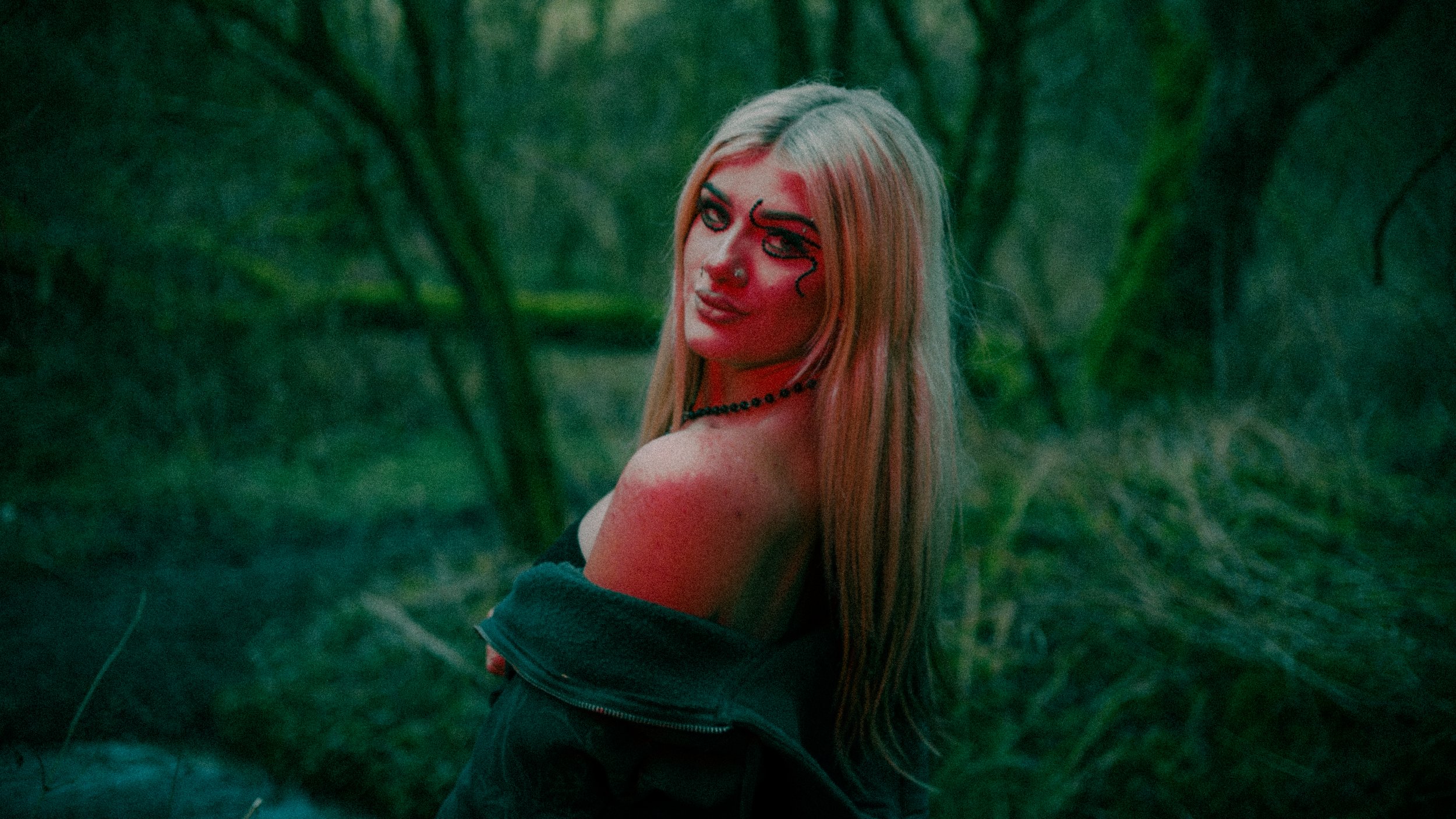 A young woman with long blonde hair and tribal-style makeup, wearing a black jacket off her shoulder, standing in a wooded area with green trees in the background at dusk or nighttime.