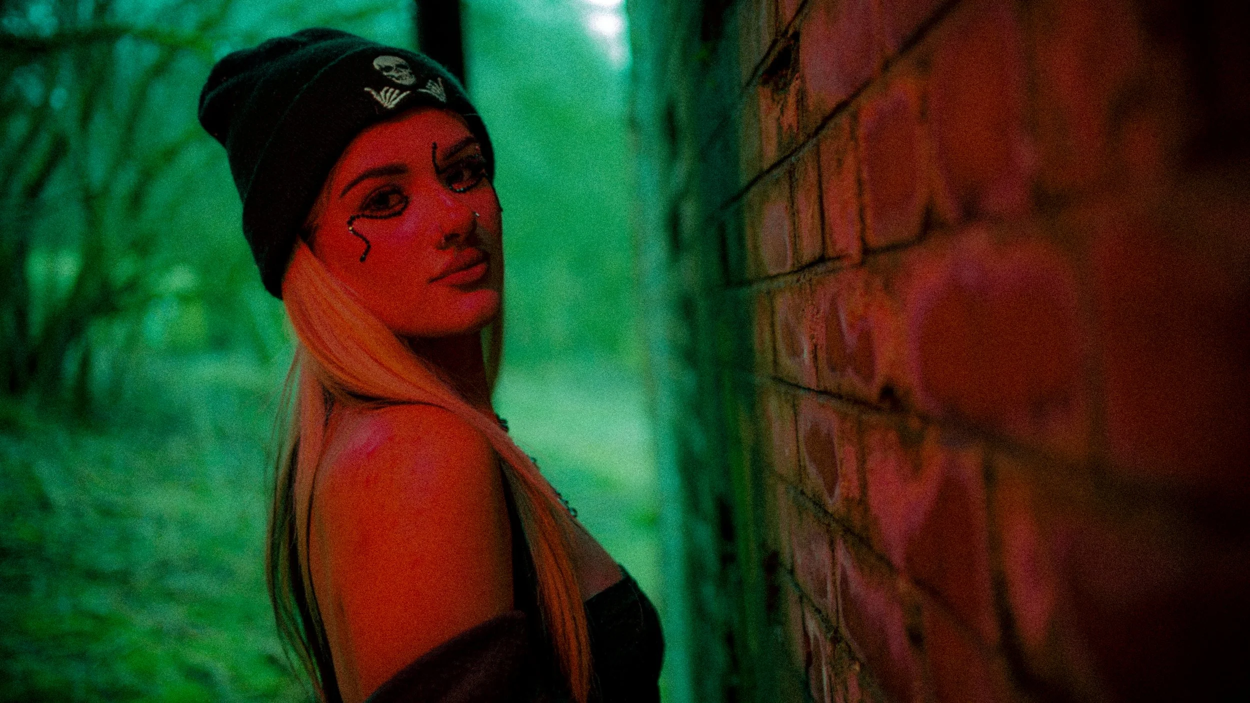 A woman with long blonde hair wearing a black beanie with a skull patch, standing against a brick wall outdoors in a wooded area, with colorful lighting.