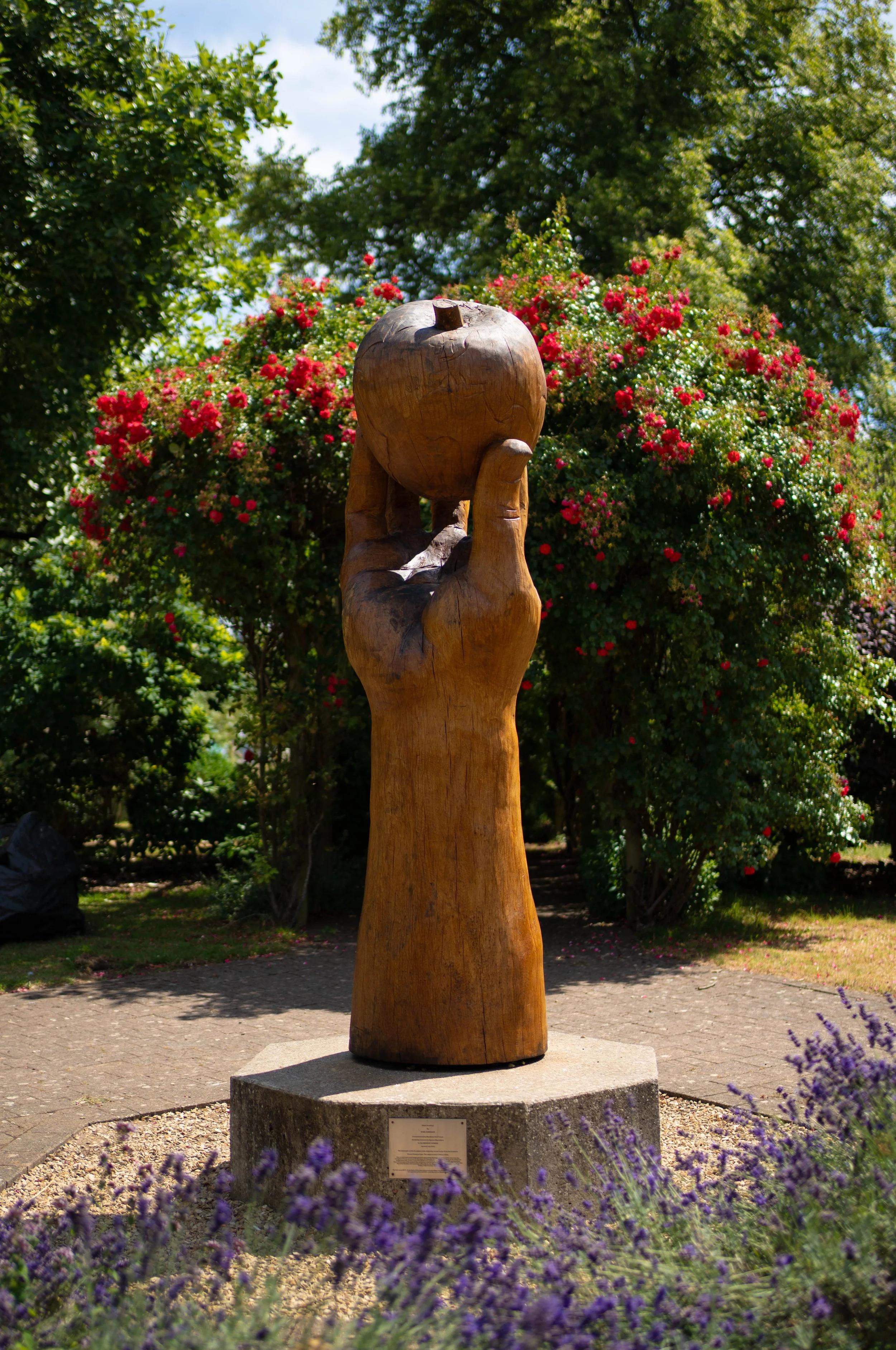 Wooden sculpture of a hand holding an apple, set outdoors in a garden with pink flowers and green trees in the background.