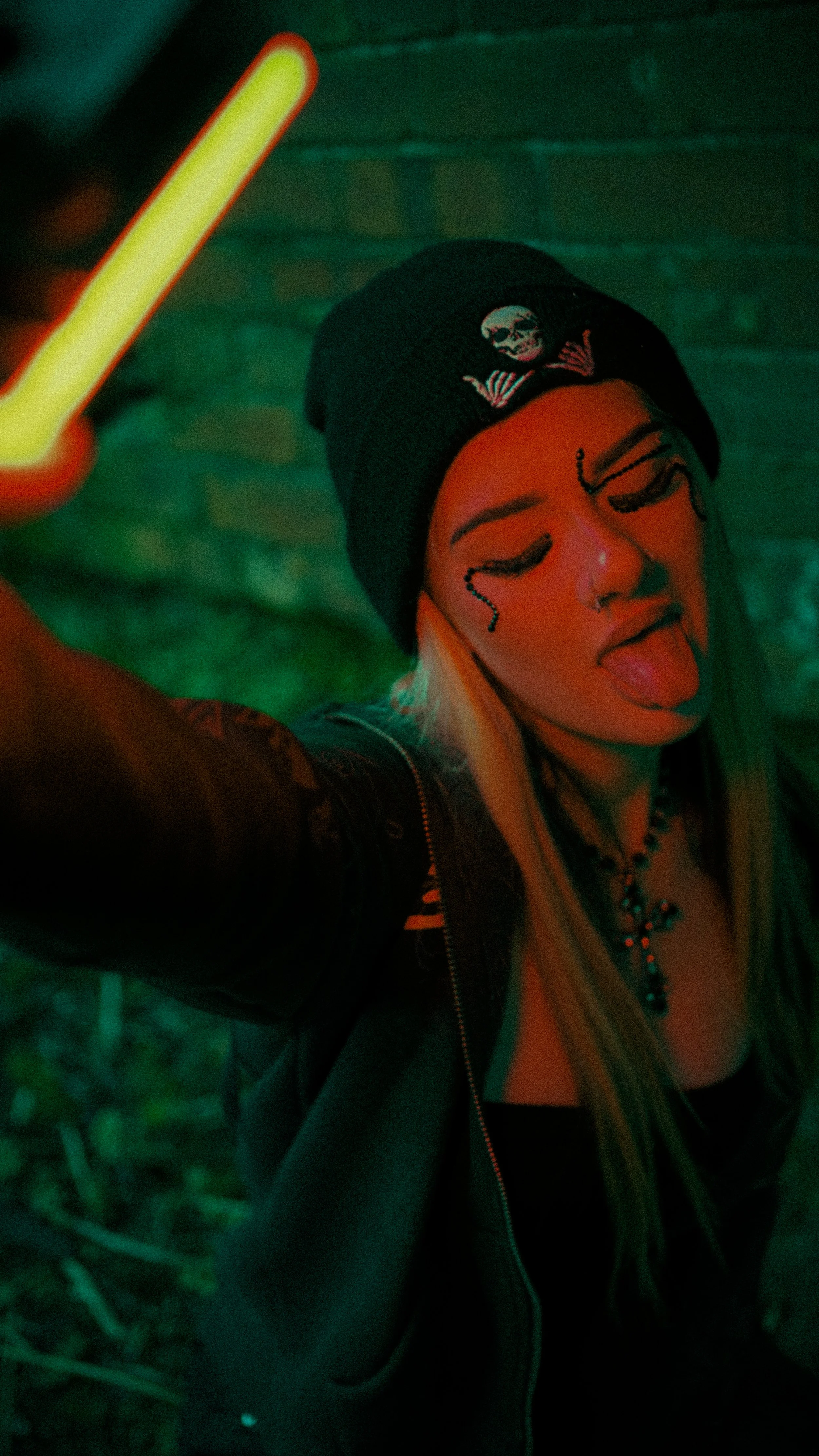 A young woman with face makeup, wearing a black beanie with a skull design, takes a selfie with her tongue out, holding a glow stick. She has long blonde hair and is dressed in dark clothing with layered necklaces, against a dark and green-illuminate