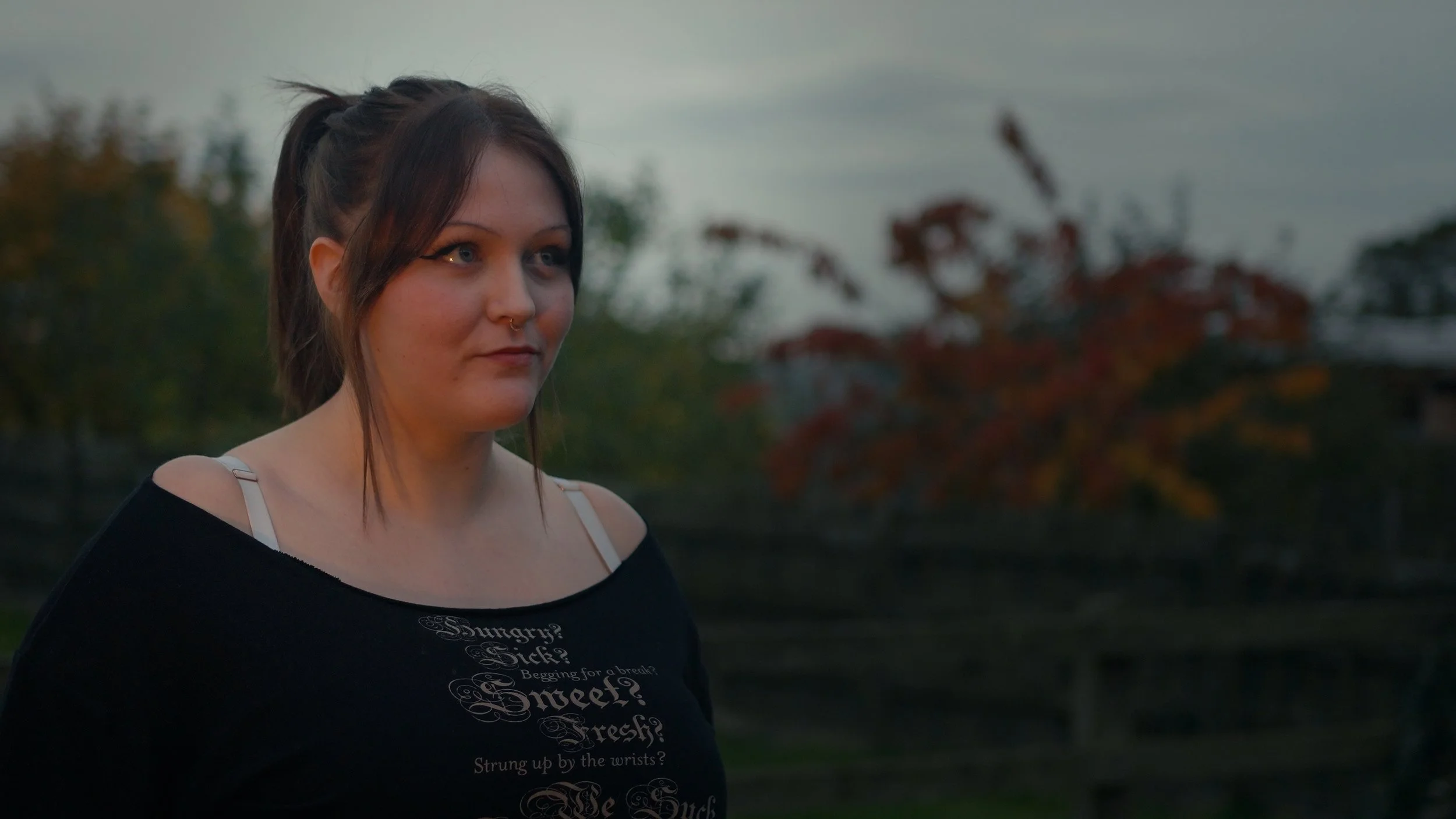 A woman with dark hair in a ponytail, wearing a black off-shoulder top with writing, standing outdoors at dusk with trees and a cloudy sky in the background.