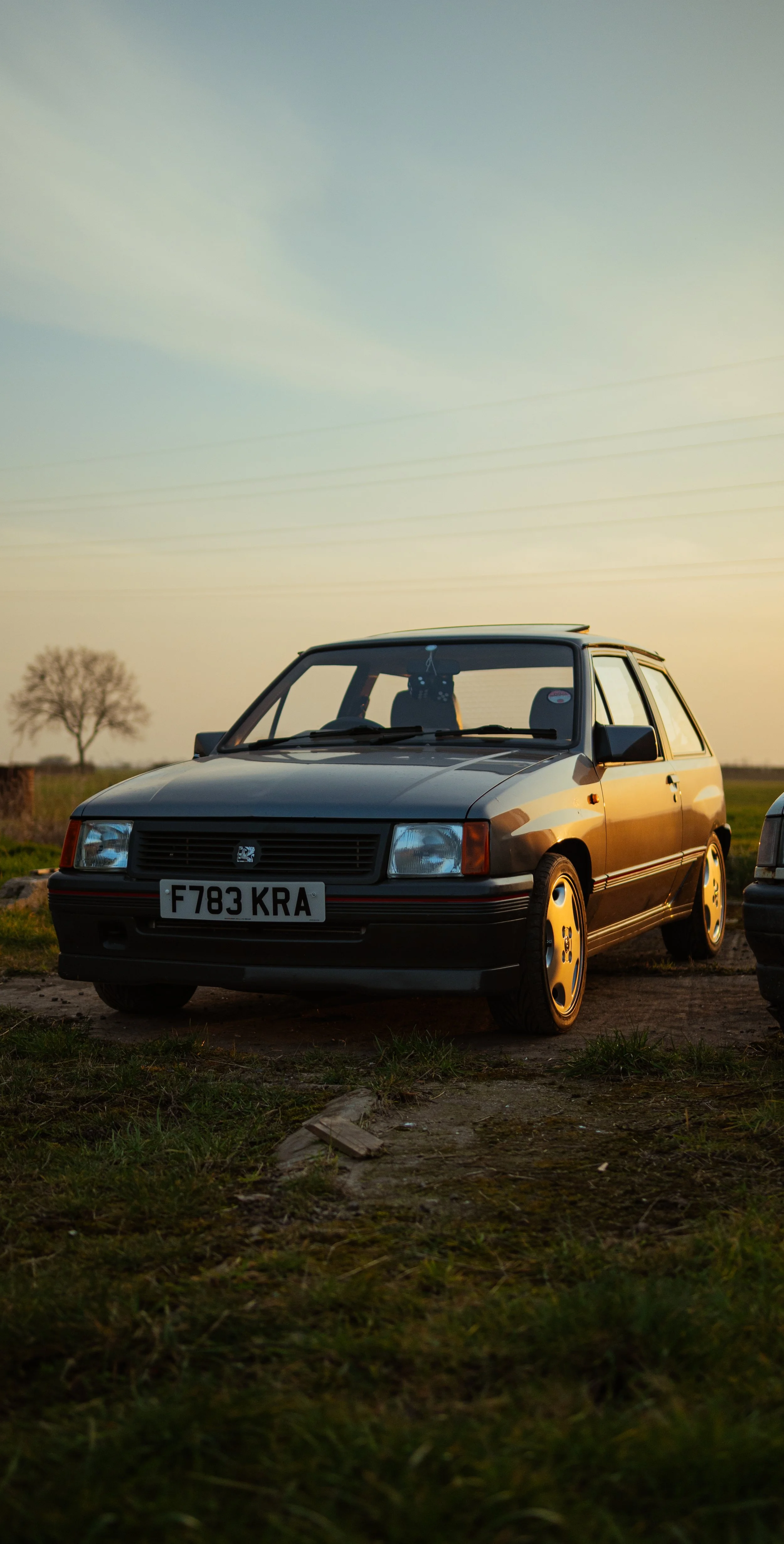 An older model black hatchback car parked outdoors on a dirt patch during sunset with a tree and open field in the background.