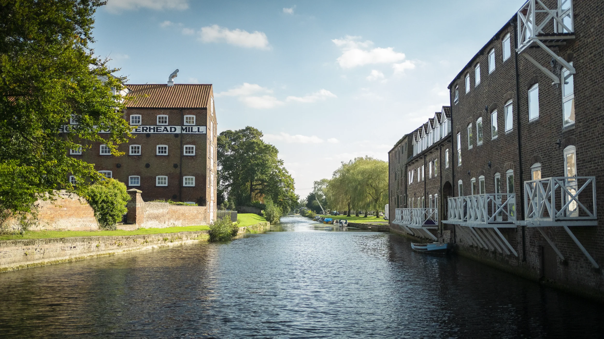 A peaceful riverside scene with brick buildings, some with white balconies, alongside a waterway with boats docked, green trees, and a partly cloudy sky.