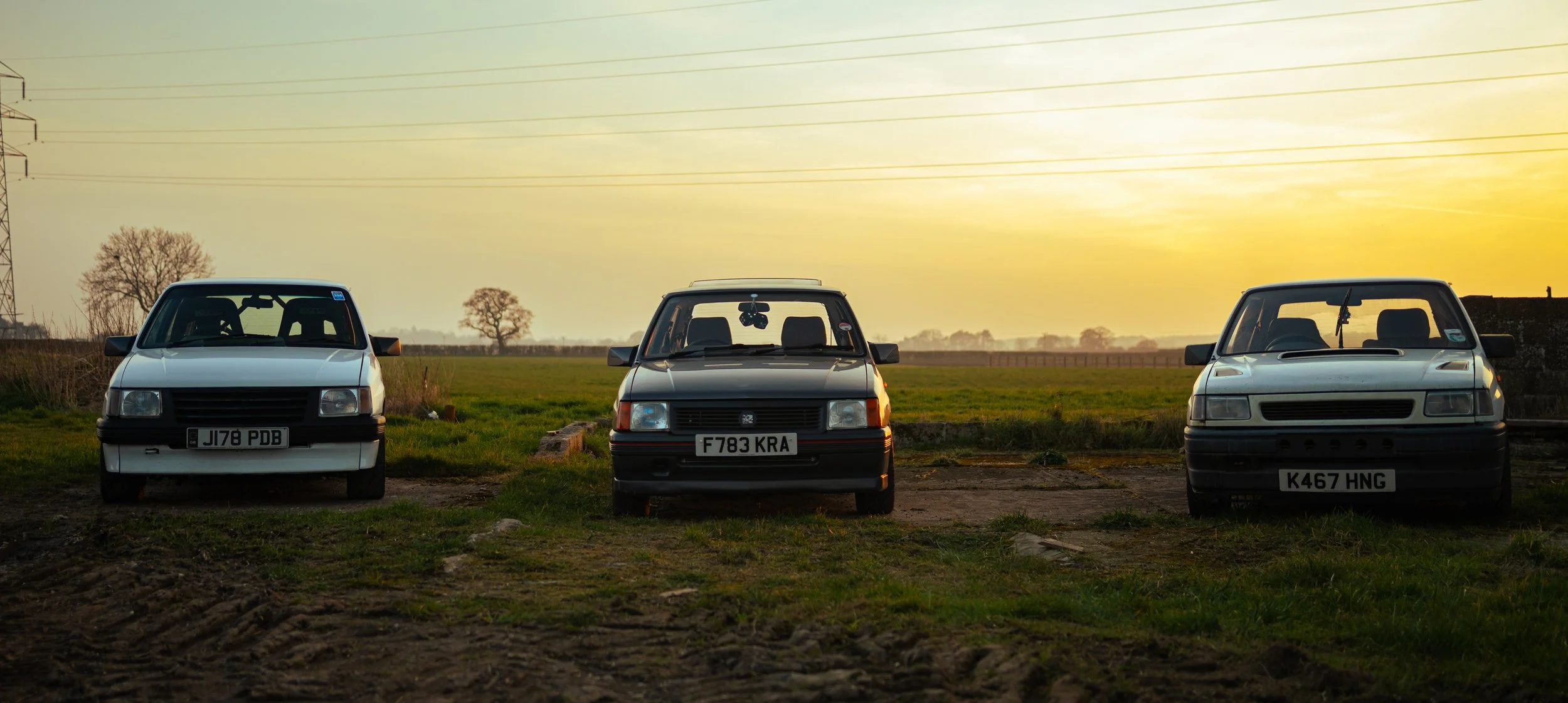Three vintage cars parked on grass field during sunset with trees and power lines in the background.