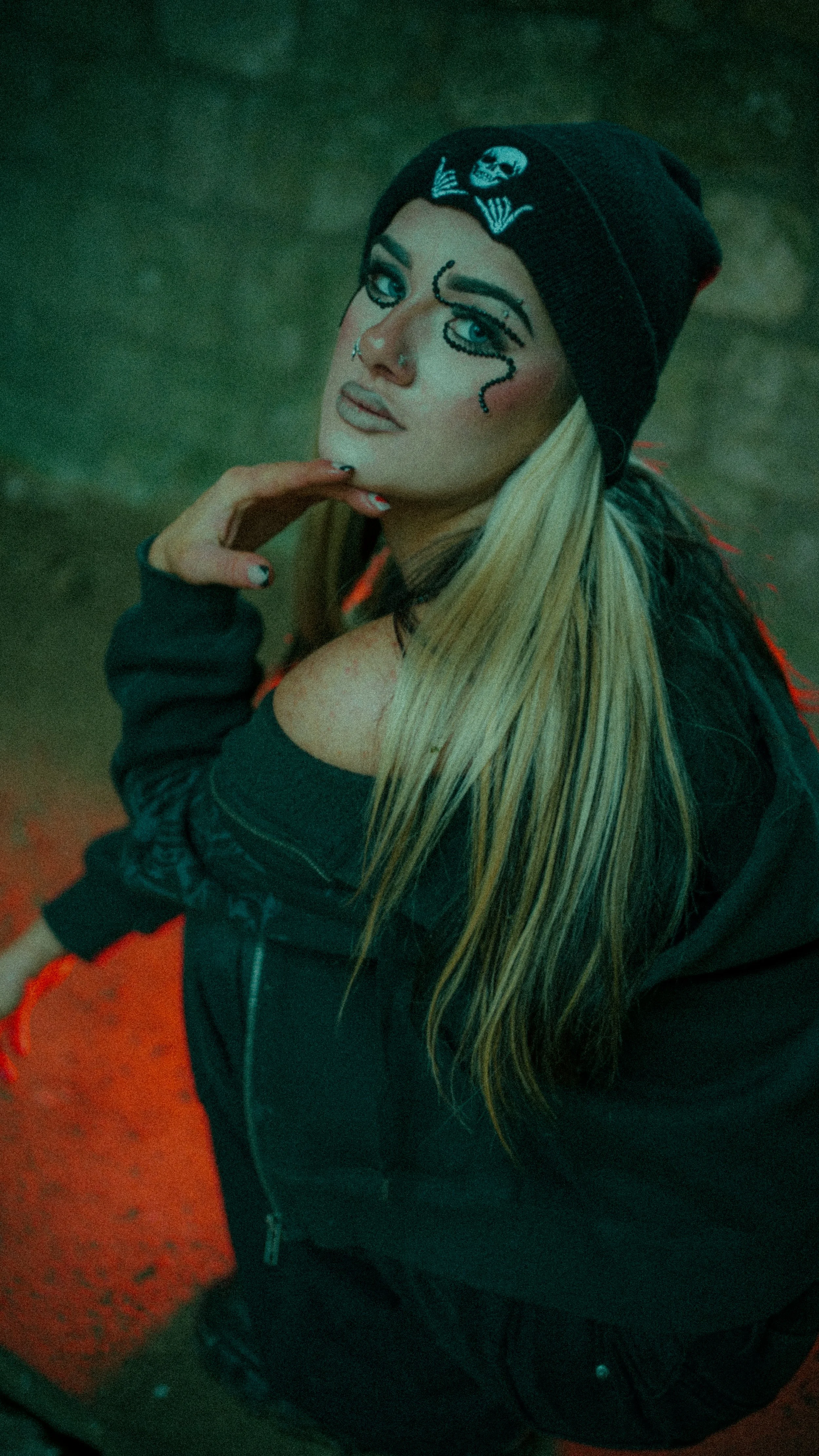 Young woman with long blonde hair, wearing a black beanie with a skull patch, a black jacket, and a dark top, standing in a forest at dusk.