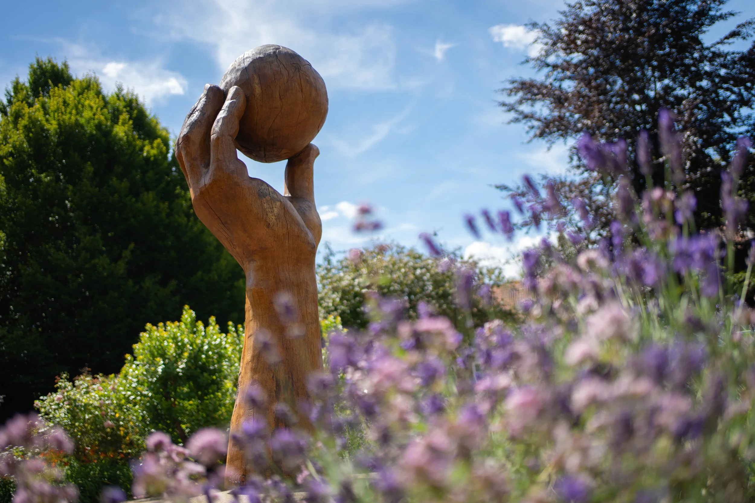 A wooden sculpture of a hand holding a sphere is situated outdoors surrounded by blooming flowers and trees under a blue sky with scattered clouds.