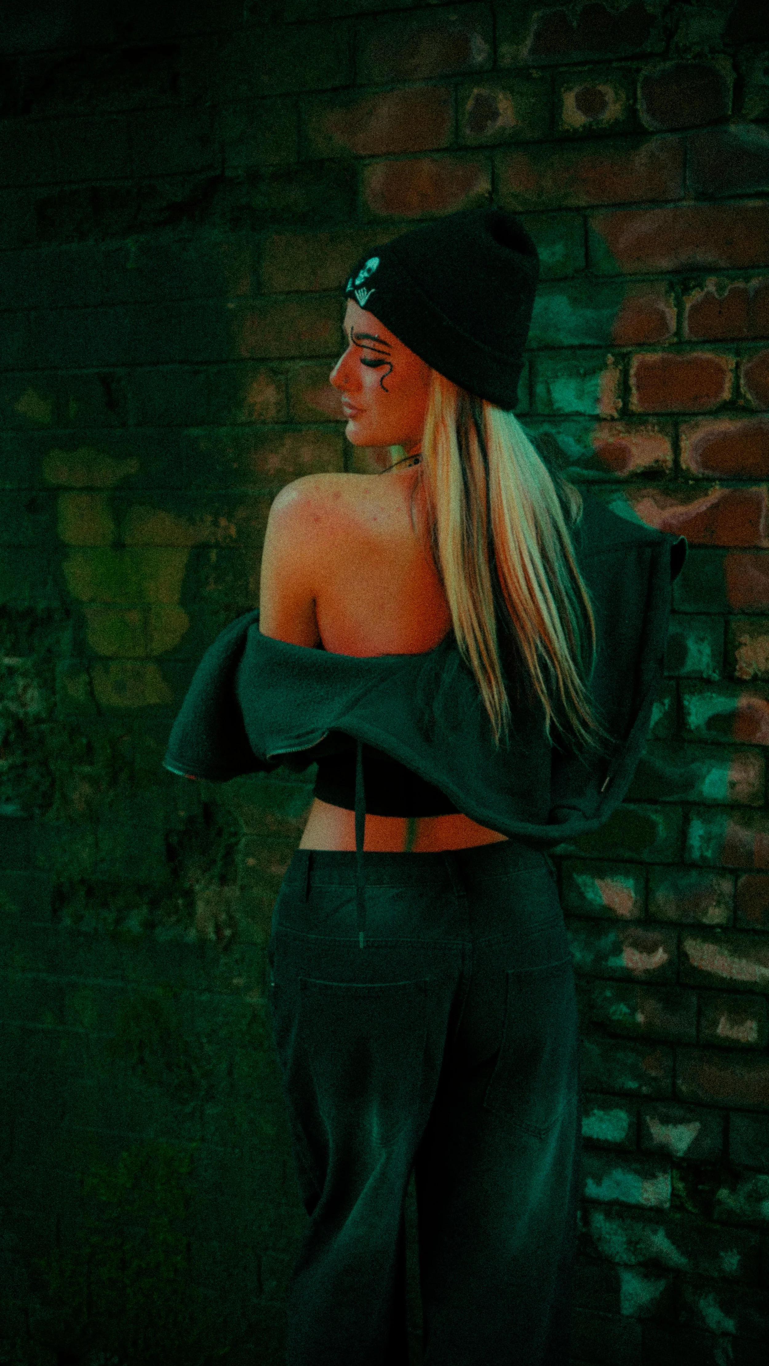 A woman with long blonde hair wearing a black beanie, off-shoulder jacket, and dark high-waisted jeans, standing against a brick wall, turned to the side with her eyes closed.