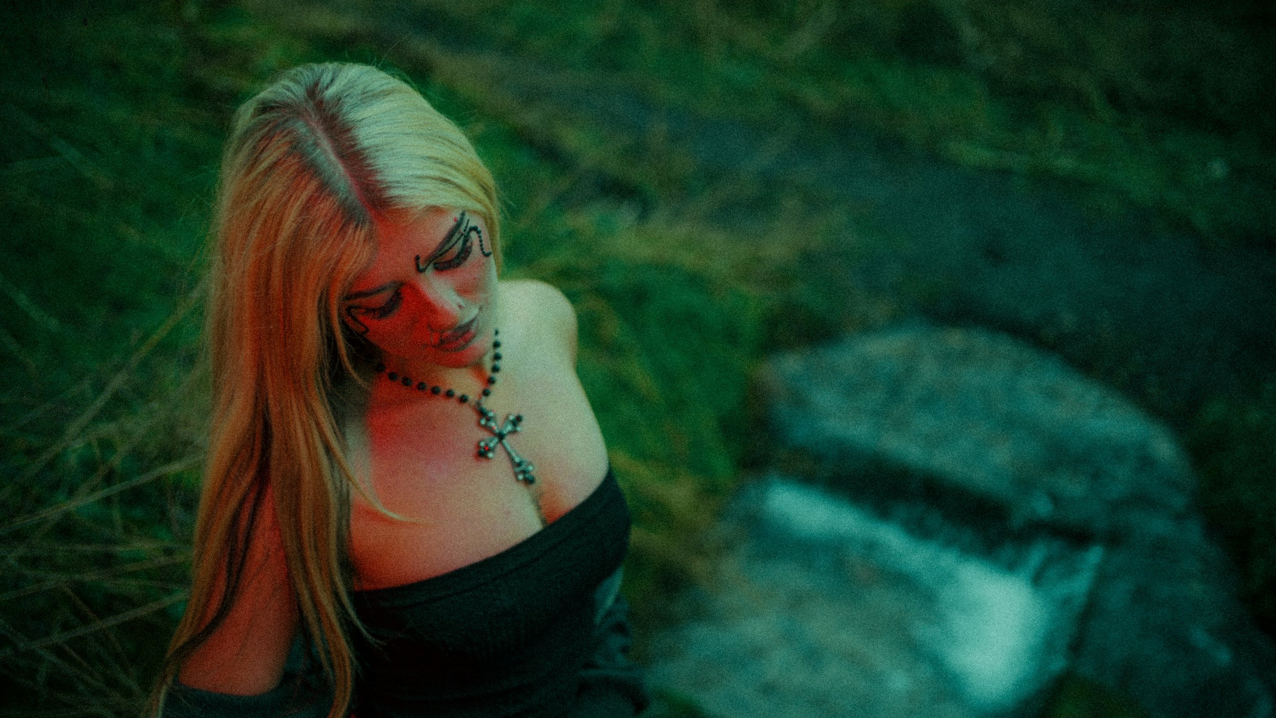 A woman with long blonde hair, wearing glasses and a black strapless top, stands outdoors near a stream with rocks and greenery, illuminated by green and red lighting, looking down.