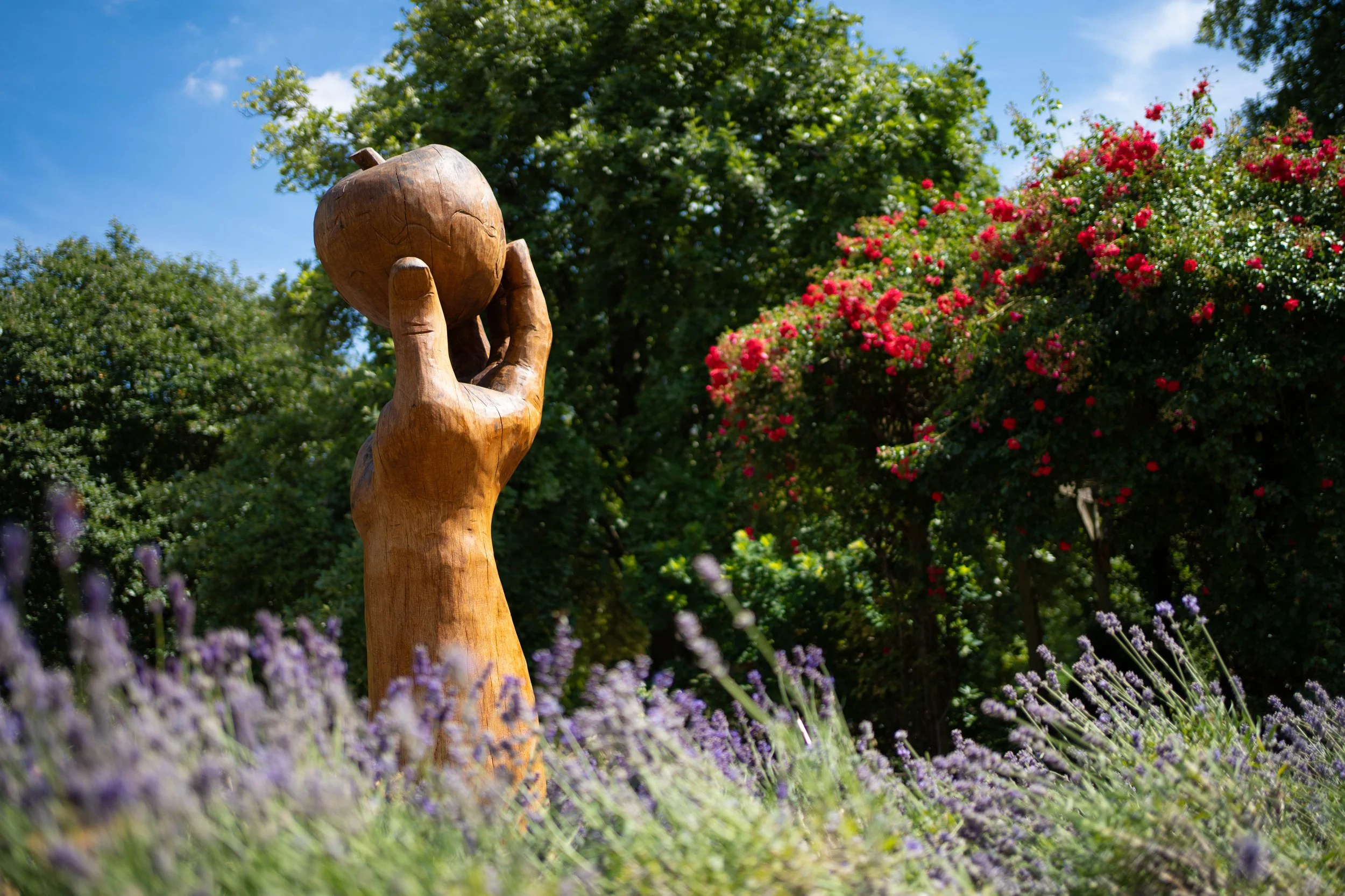A wooden sculpture of a hand holding an apple, set outdoors among green trees and blooming purple and red flowers on a sunny day.