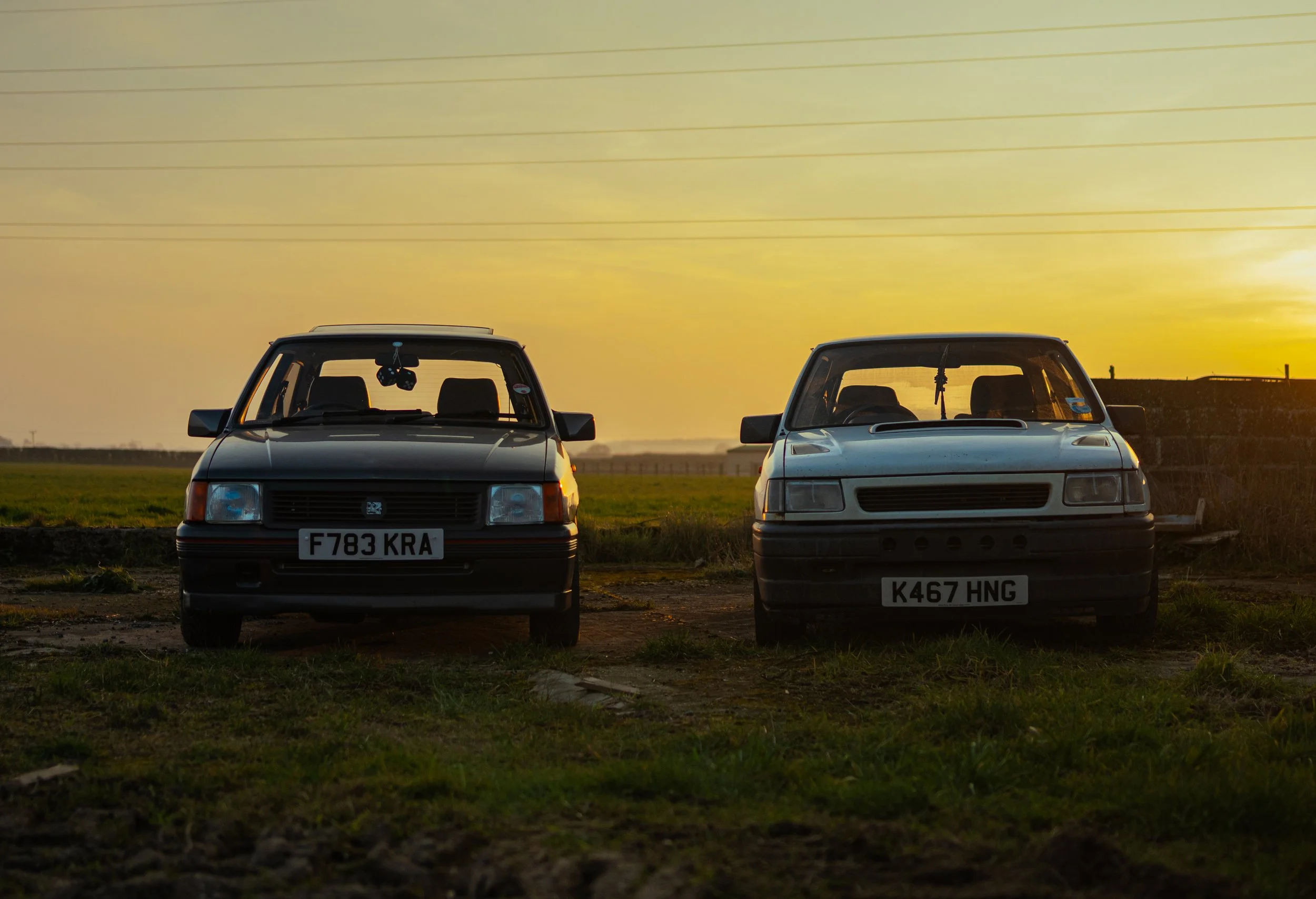 Two classic cars parked on a grassy field at sunset, with the sky in warm tones and power lines overhead.