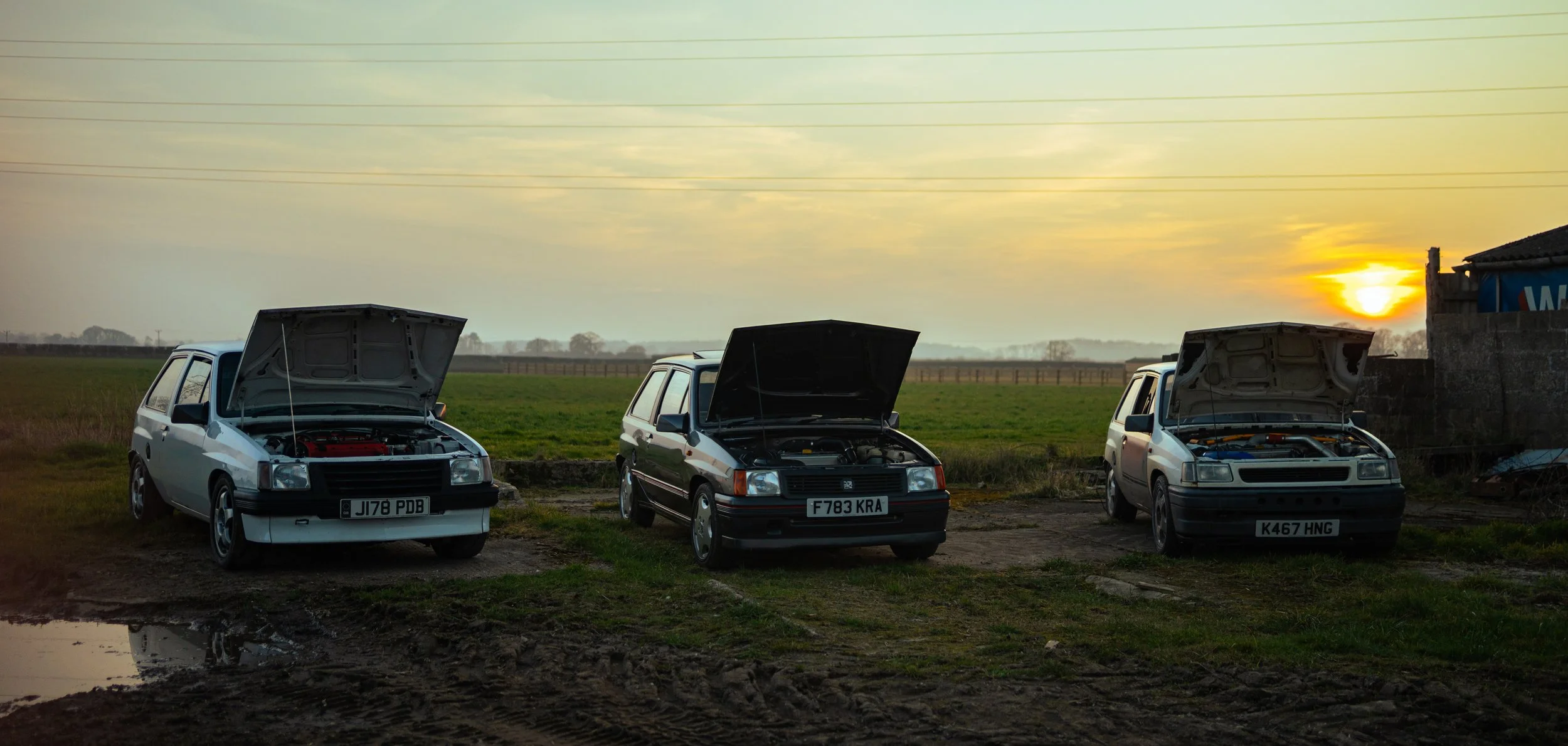 Three vintage cars with open hoods parked on a grassy area near a farm field at sunset, with a building to the right.